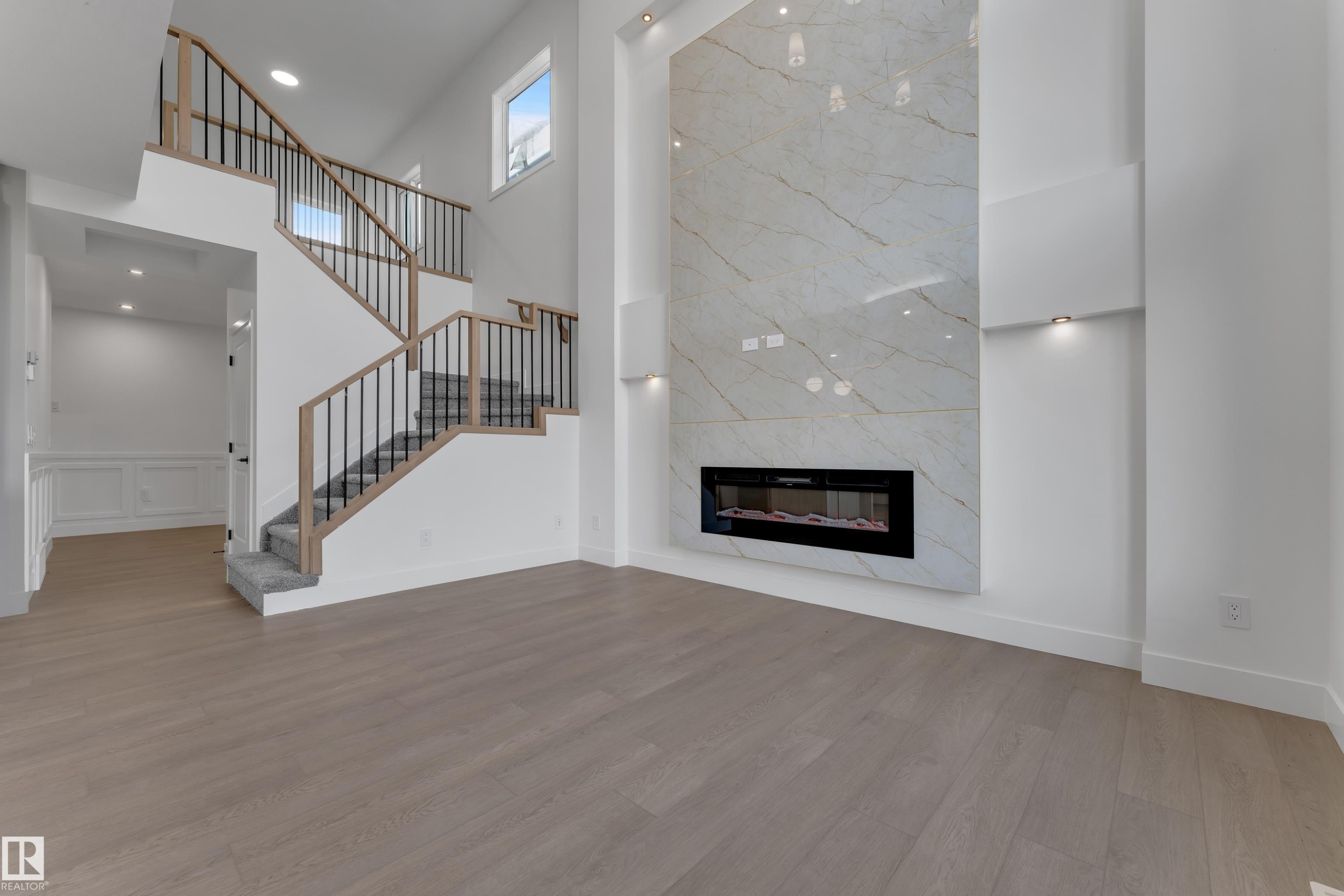 Two-story living area with wood-finish flooring, a contemporary fireplace feature wall, recessed lighting, and a staircase with wood handrails and black metal spindles - 247 Linden Loop, Leduc, AB