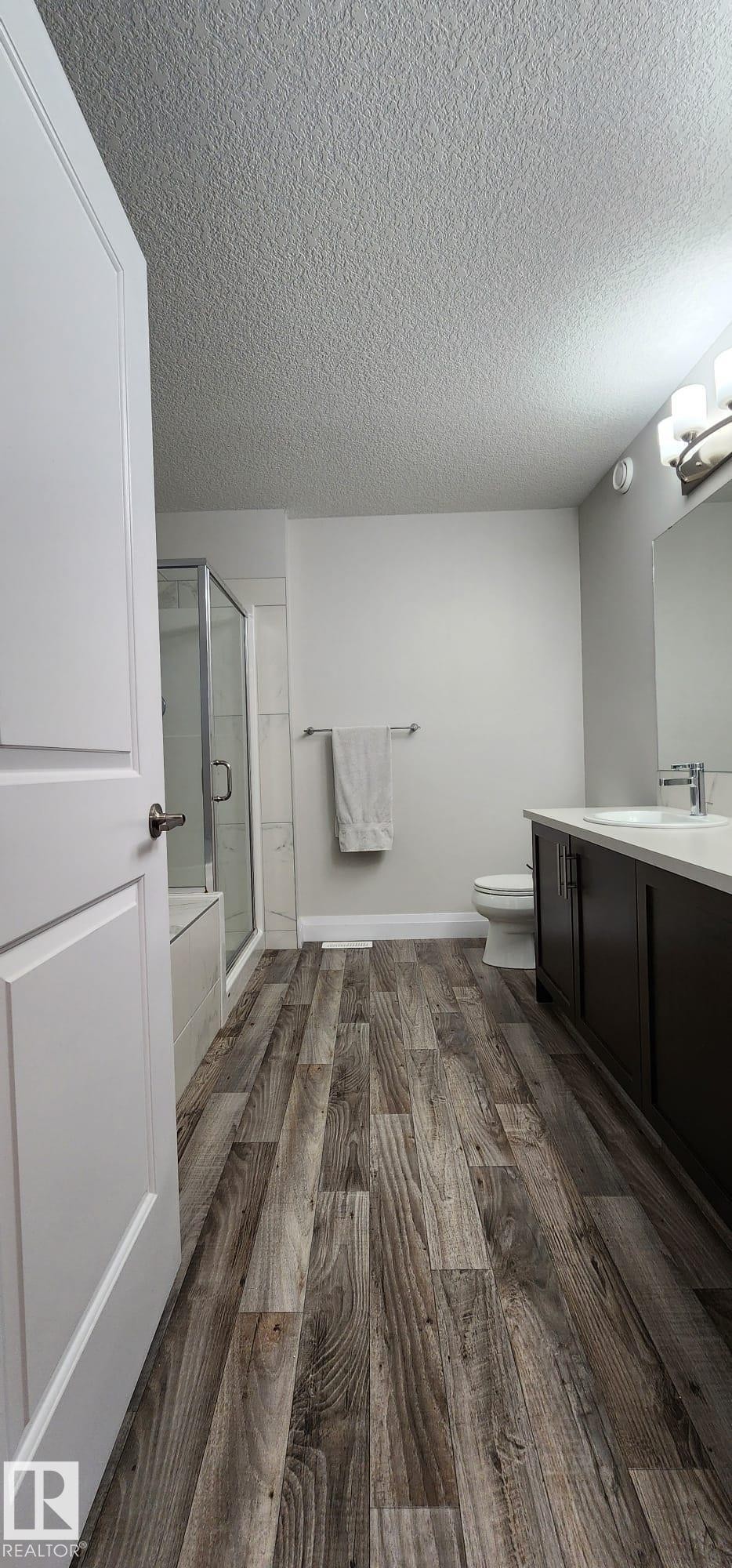 Wood-finish flooring throughout, dark wood-finish vanity with white countertop, recessed lighting, white tub with glass enclosure, and white walls - 3779 Cherry Loop Sw, Edmonton, AB