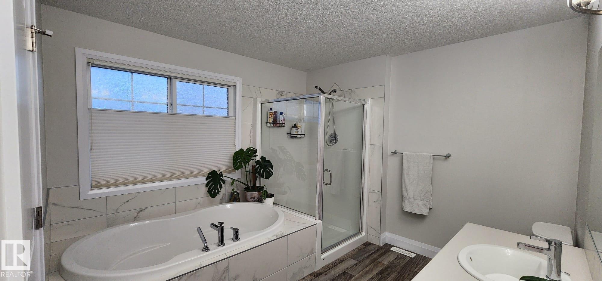 Soaking tub with a tiled surround, separate glass-enclosed shower, dual-pane window, and wood-finish flooring - 3779 Cherry Loop Sw, Edmonton, AB