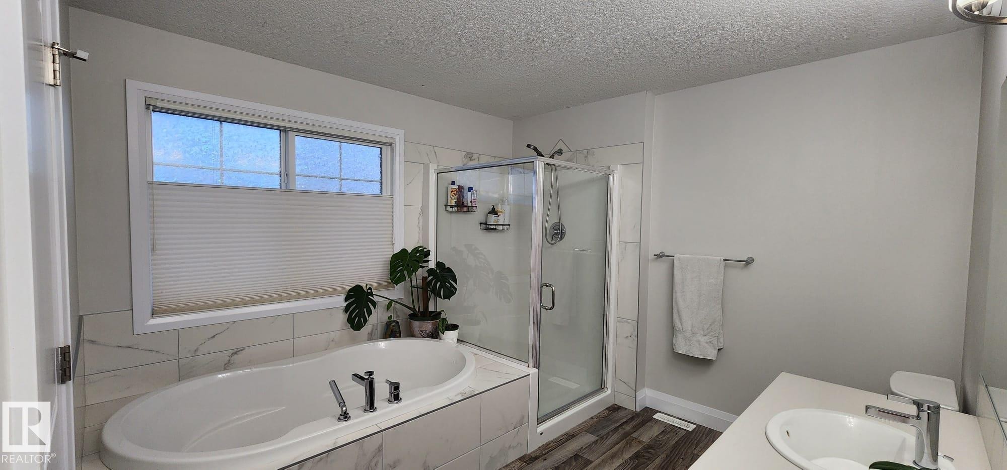 Oval soaking tub with deck-mounted fixtures, glass-enclosed shower, white vanity with integrated sink, and wood-finish flooring - 3779 Cherry Loop Sw, Edmonton, AB