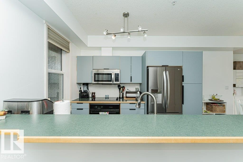 Well-appointed kitchen featuring light blue cabinetry, stainless steel appliances, a modern track lighting fixture, a stainless steel sink with a gooseneck faucet, and a breakfast bar with a speckled green countertop - #202 9503 101 Av Nw, Edmonton, AB