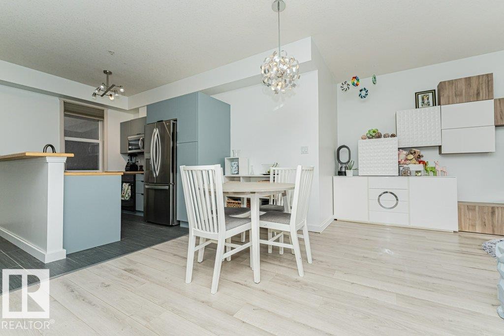 Open-concept living area with light wood-finish flooring, a contemporary chandelier, recessed lighting, and a kitchen featuring stainless steel appliances and a breakfast bar with a wood-finish countertop - #202 9503 101 Av Nw, Edmonton, AB
