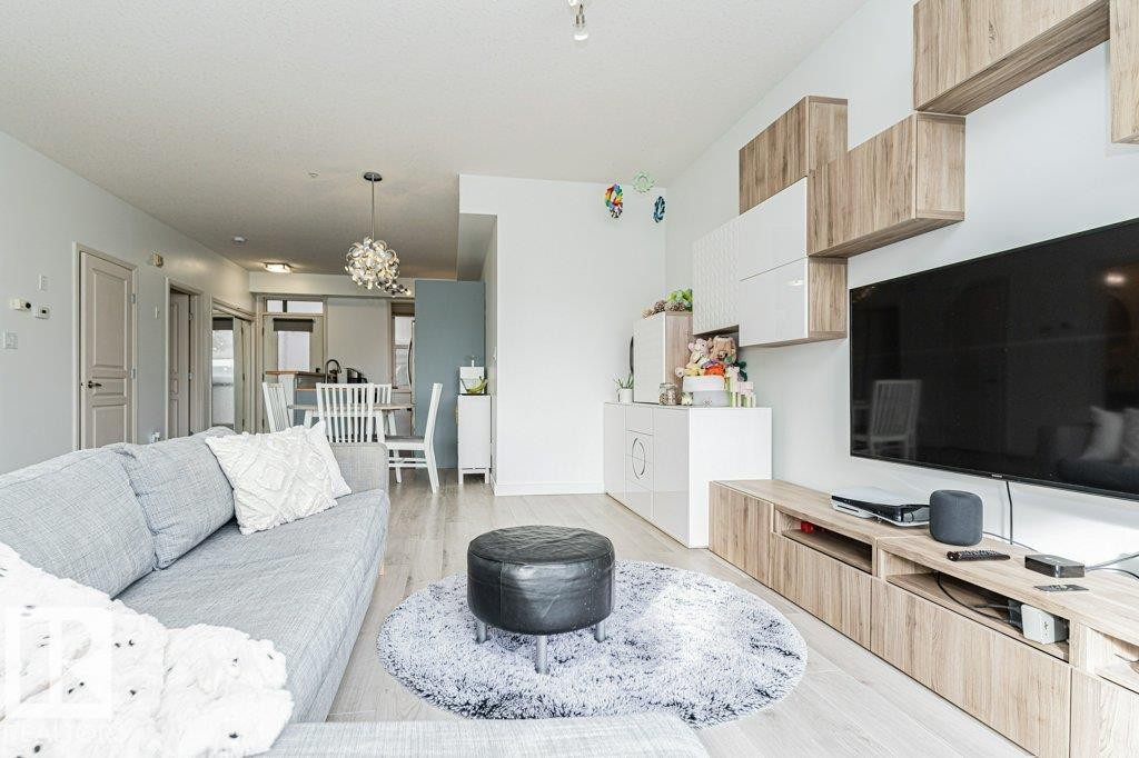 Light wood-finish flooring throughout, integrated media console with wood-finish cabinetry, modern wall-mounted shelving, contemporary dining area pendant lighting, and a kitchen with white cabinetry - #202 9503 101 Av Nw, Edmonton, AB