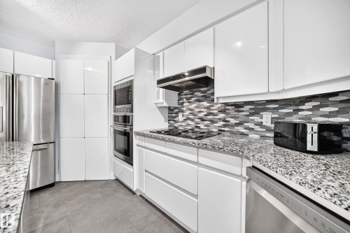 White gloss cabinetry, stainless steel appliances, speckled stone countertops, mosaic tile backsplash, and built-in double ovens - 5830 181 Street Nw, Edmonton, AB 