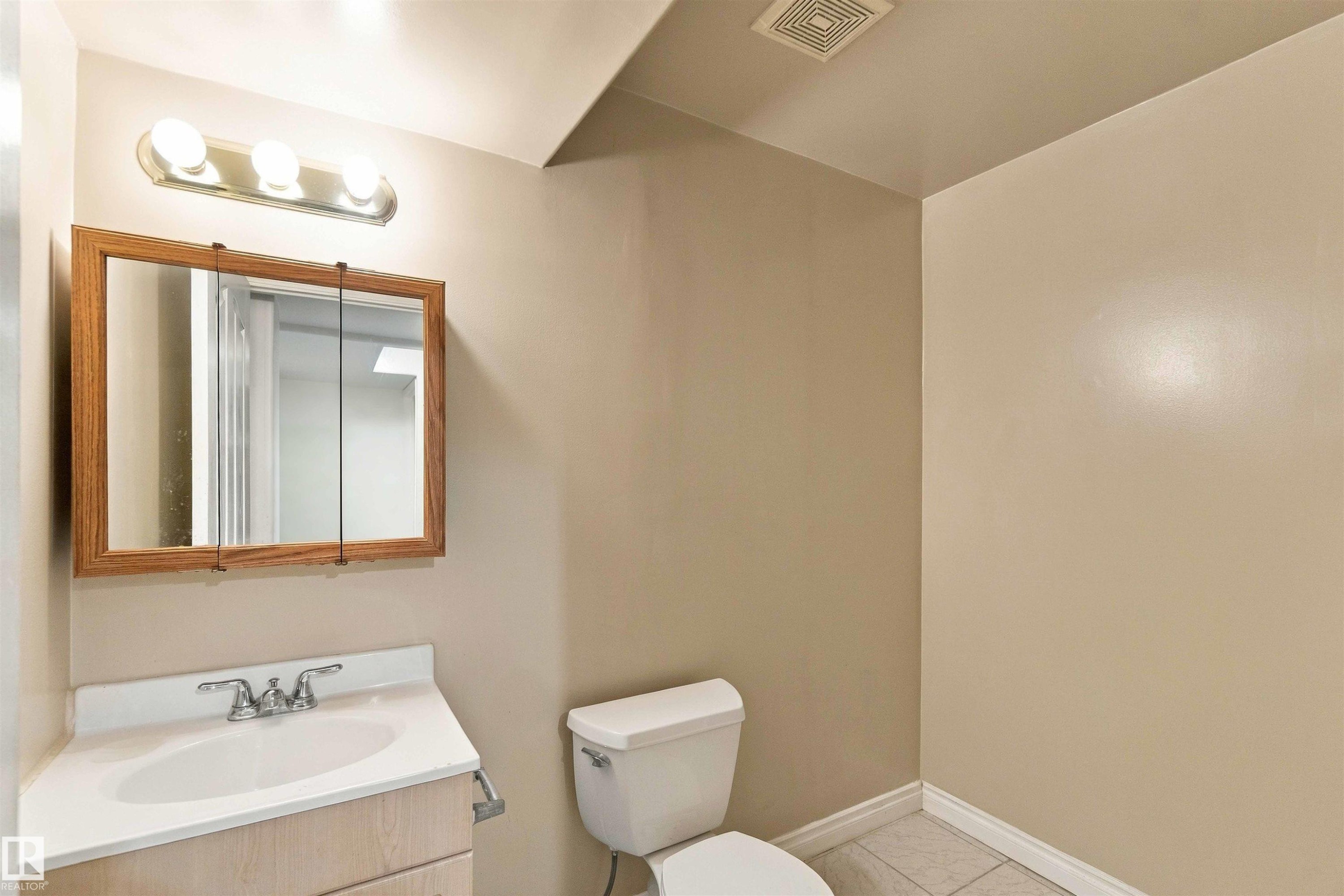 Wall-mounted light fixture with three bulbs, wood-frame medicine cabinet, vanity with integrated sink, chrome faucet, and tile flooring - 3524 Mclean Crescent Sw, Edmonton, AB