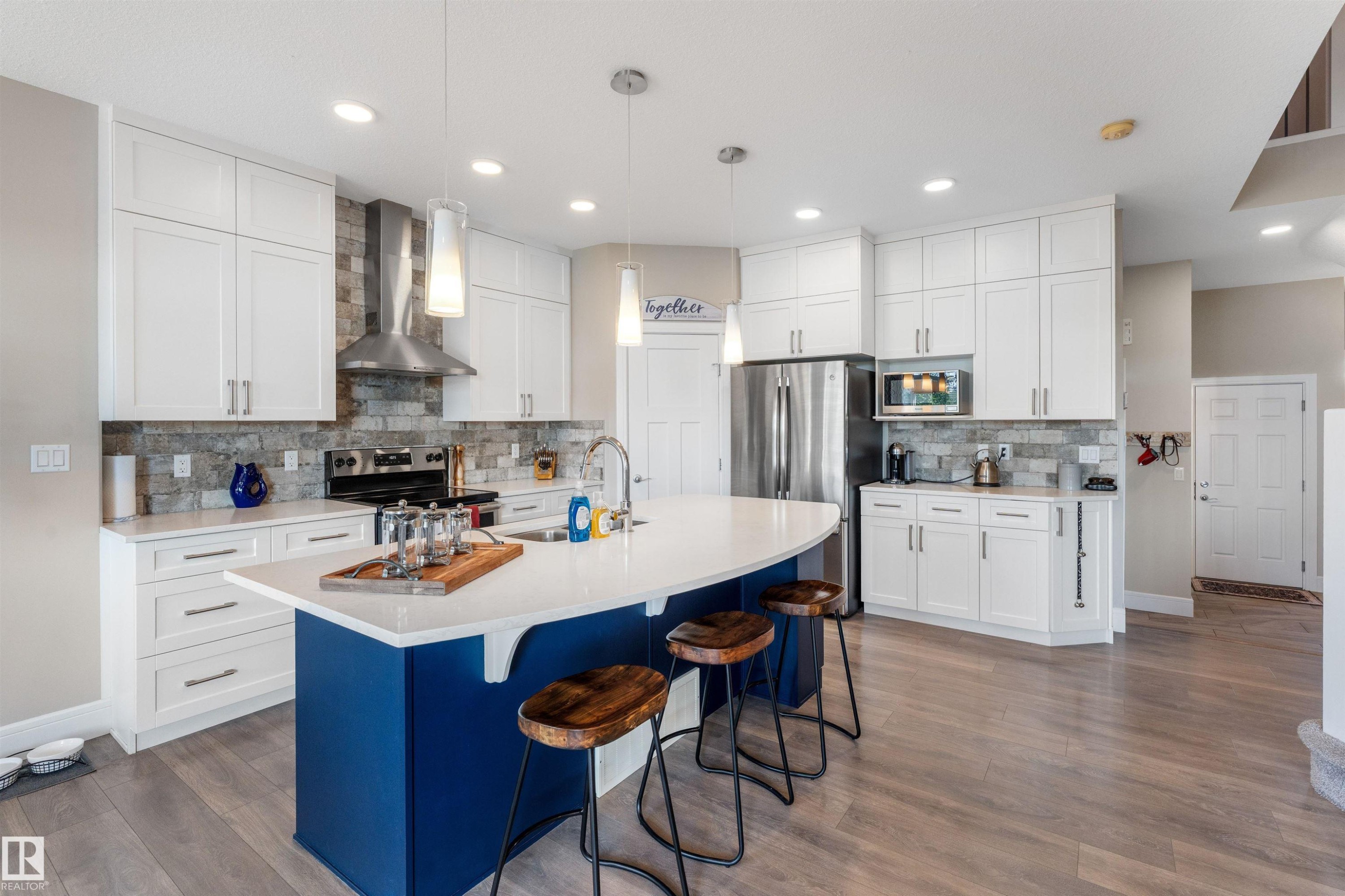 Kitchen featuring a large central island with a curved countertop, white cabinetry, integrated stainless steel appliances, wood-finish flooring, and a stone tile backsplash - 3334 Parker Loop Sw, Edmonton, AB