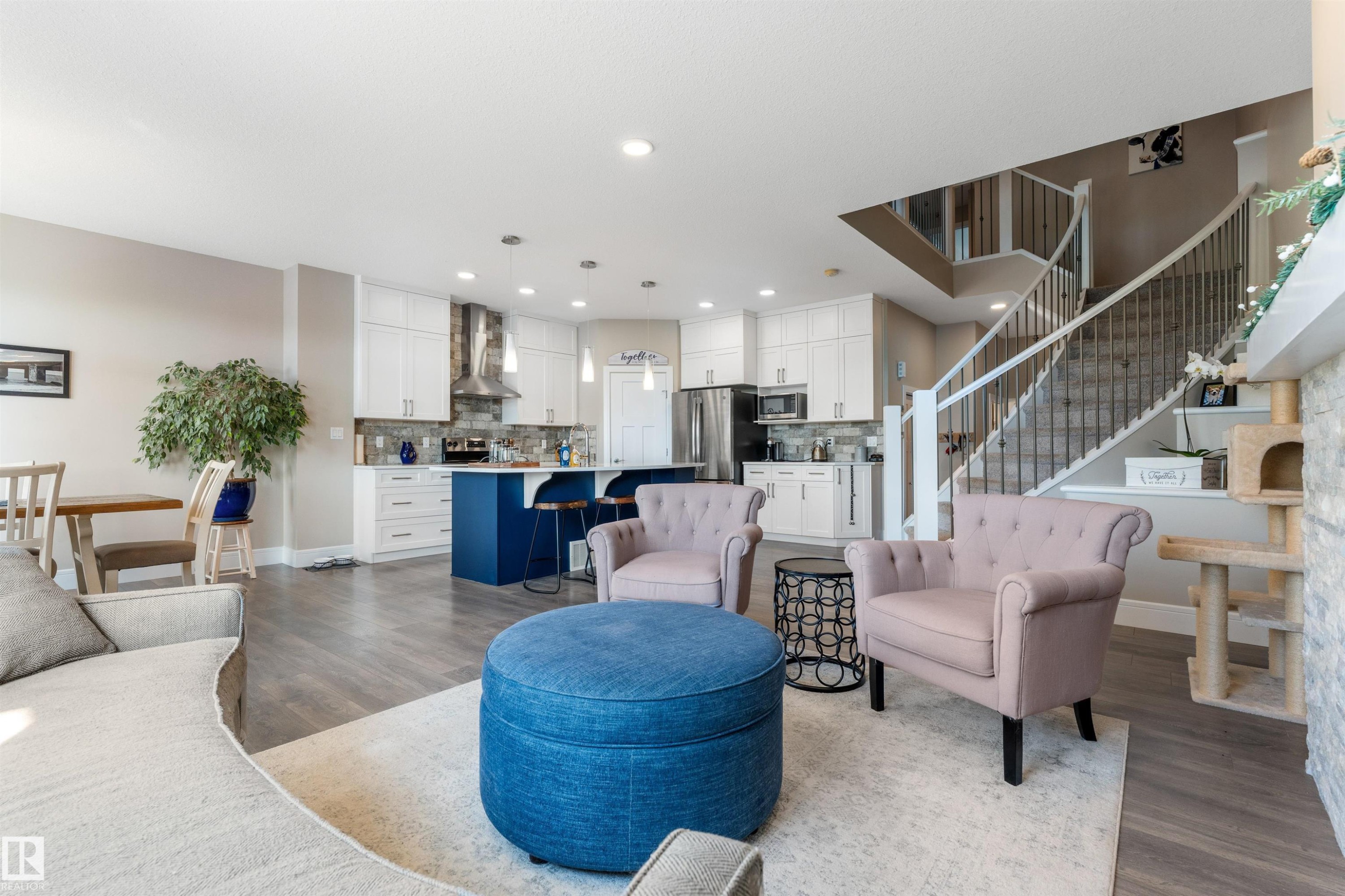 Open-concept living space featuring wood-finish flooring, a contemporary kitchen with white cabinetry and a blue island, and a prominent staircase with white risers and dark balusters - 3334 Parker Loop Sw, Edmonton, AB