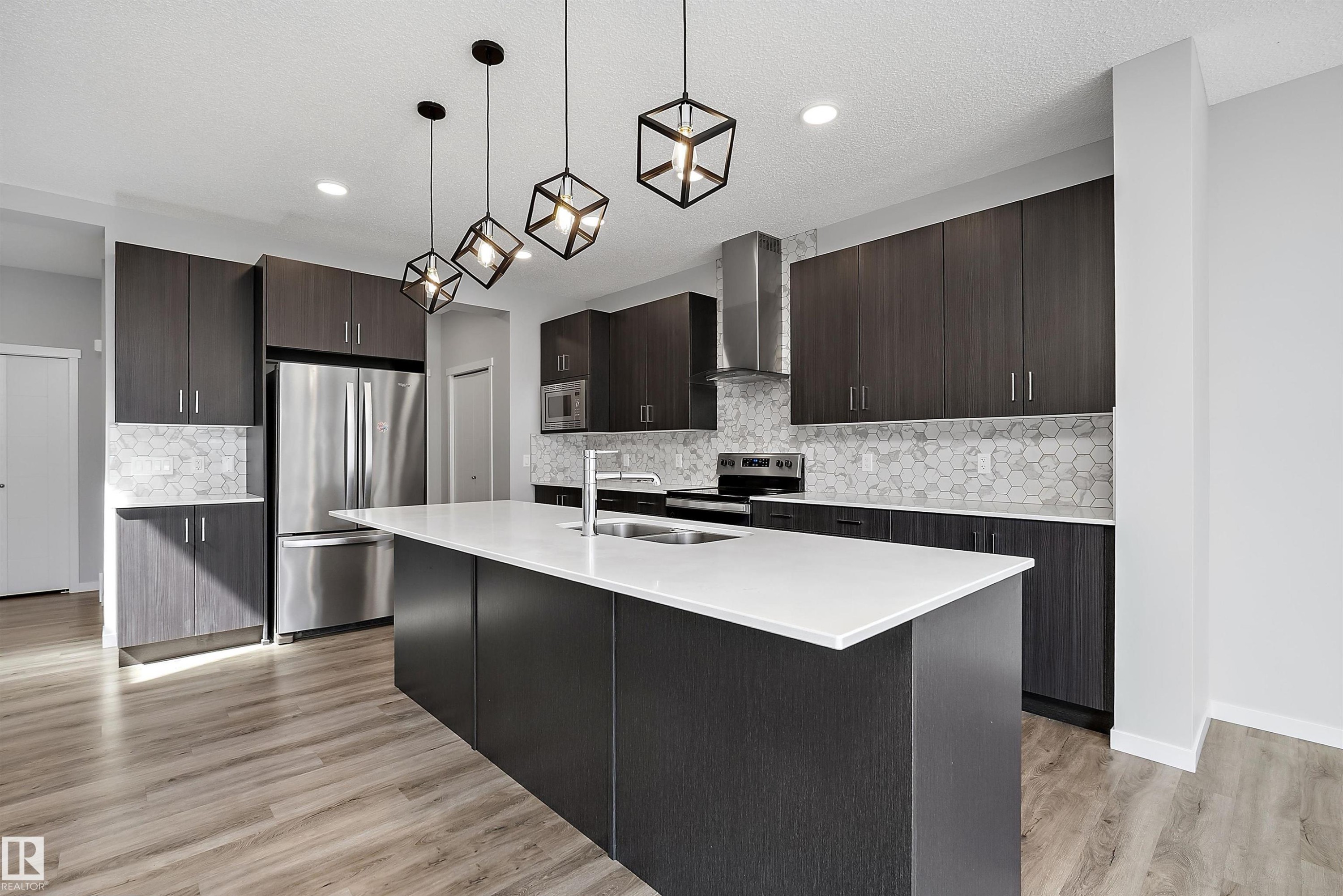 Kitchen featuring wood-finish flooring, dark wood-finish cabinetry, a large kitchen island with a white countertop, stainless steel appliances, and geometric pendant lighting - 9227 223 Street Nw, Edmonton, AB