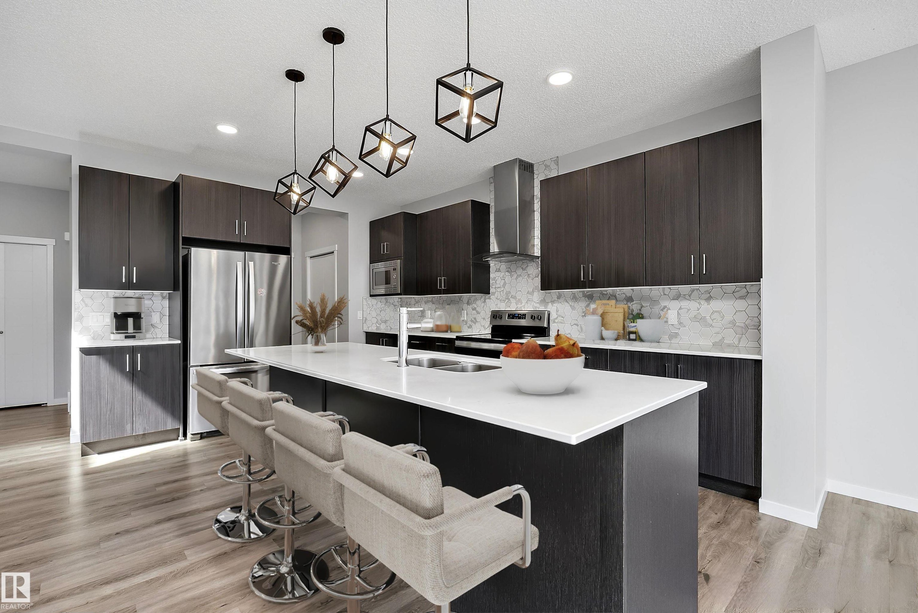 Contemporary kitchen featuring dark wood-finish cabinetry, stainless steel appliances, a white countertop island with seating, geometric pendant lighting, and light wood-finish flooring - 9227 223 Street Nw, Edmonton, AB