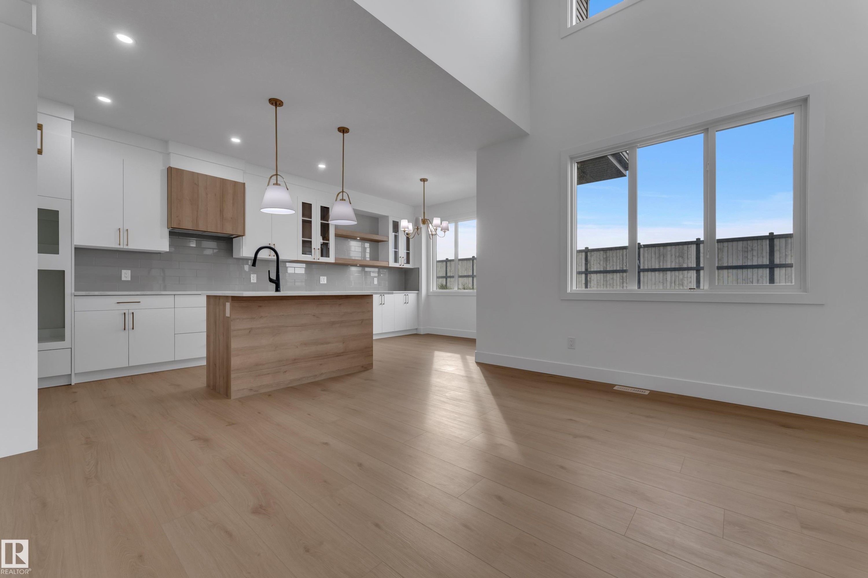 Open-concept living area featuring light wood-finish flooring, a kitchen with white cabinetry and a wood-finish island, a gray tile backsplash, and large windows - 178 Munn Way, Leduc, AB