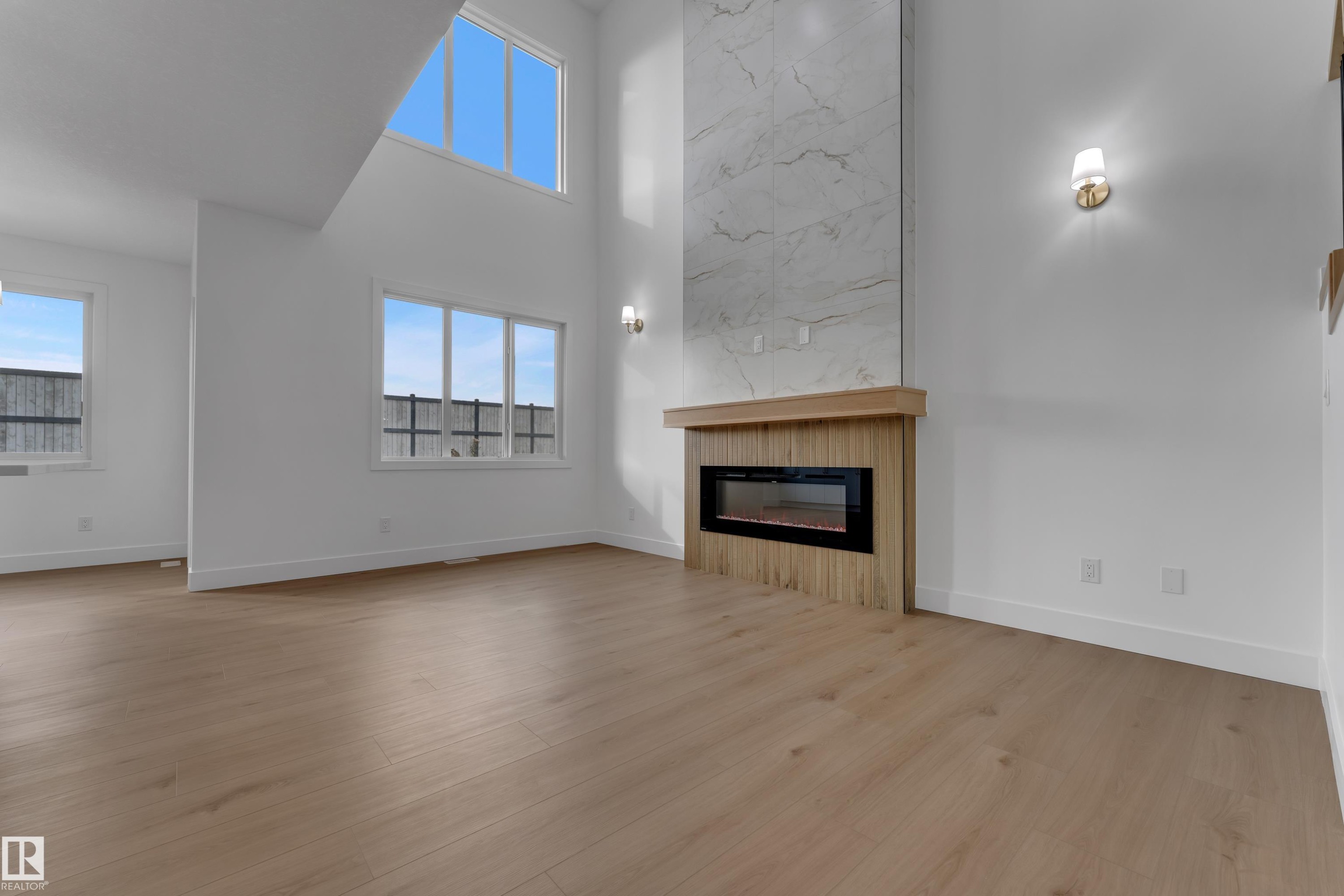 Expansive room featuring high ceilings, wood-finish flooring, an electric fireplace with a wood mantle and tile surround, and multiple windows - 178 Munn Way, Leduc, AB