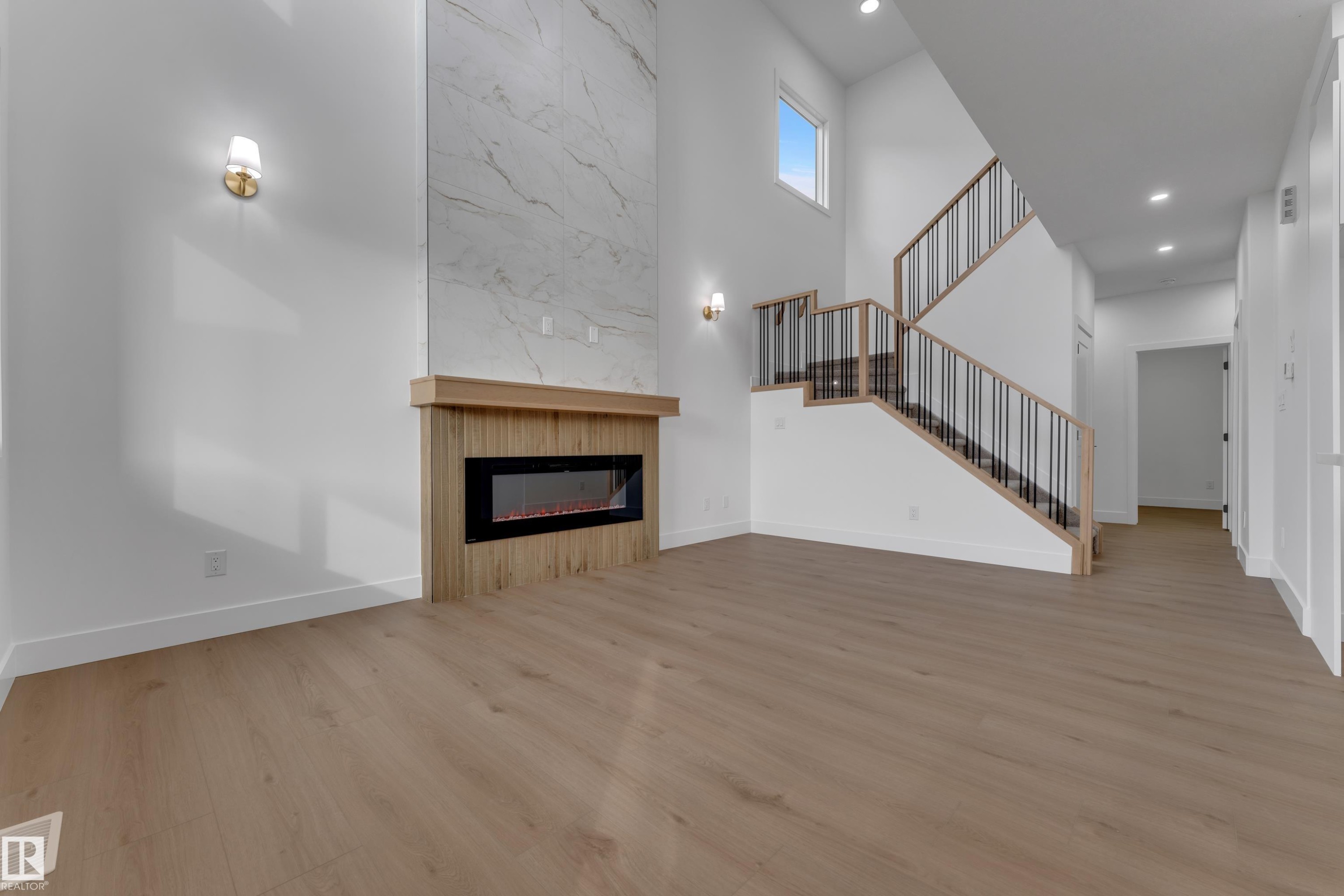 Two-story living area featuring a wood-finish electric fireplace with a floor-to-ceiling marble-look surround, light wood-finish flooring, and recessed lighting - 178 Munn Way, Leduc, AB