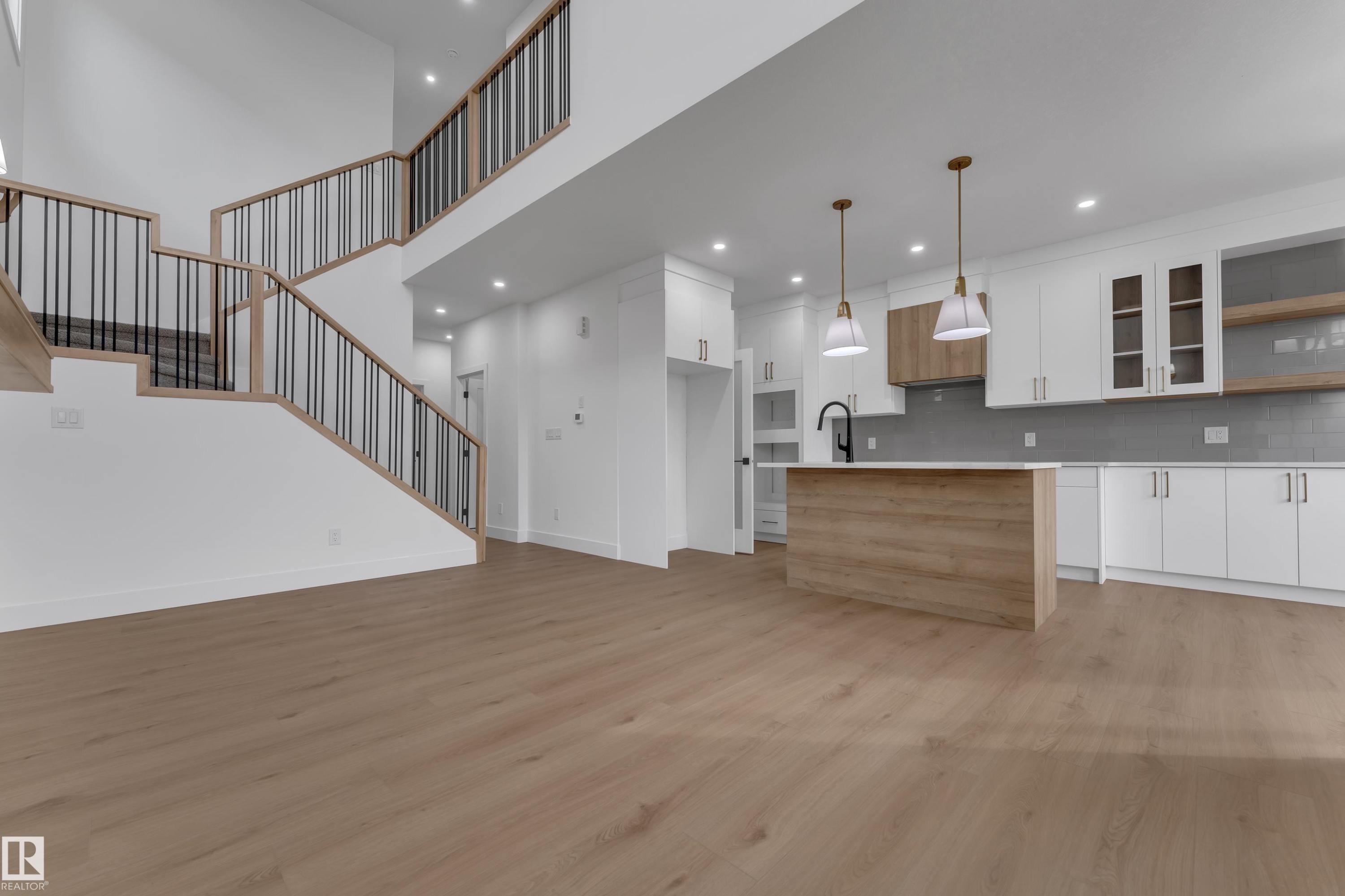 Open-concept living space featuring wood-finish flooring, a contemporary kitchen with white cabinetry, a large kitchen island, and a prominent staircase with dark metal balusters - 178 Munn Way, Leduc, AB