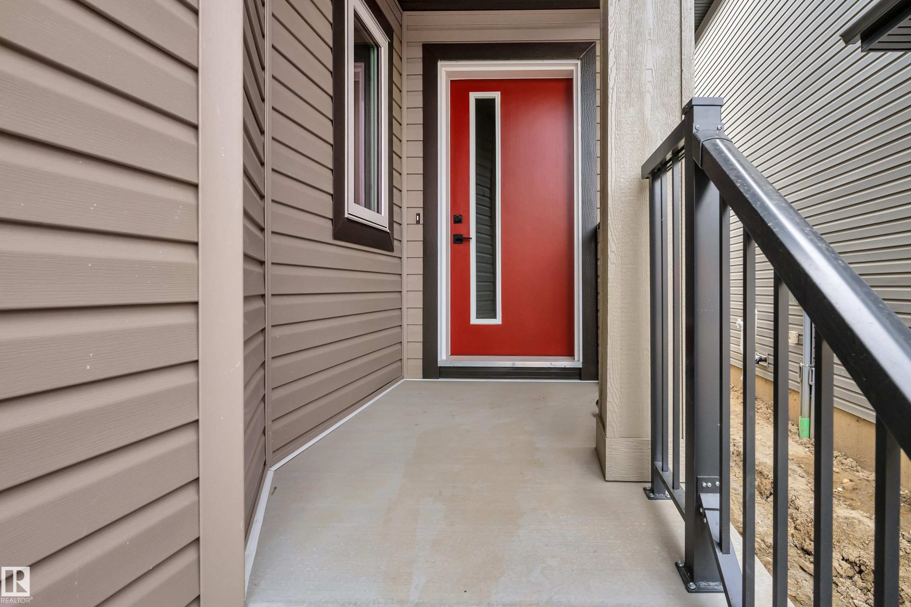 Entryway featuring a vibrant red door with vertical glass insert, light-colored concrete porch, and black metal railing - 178 Munn Way, Leduc, AB