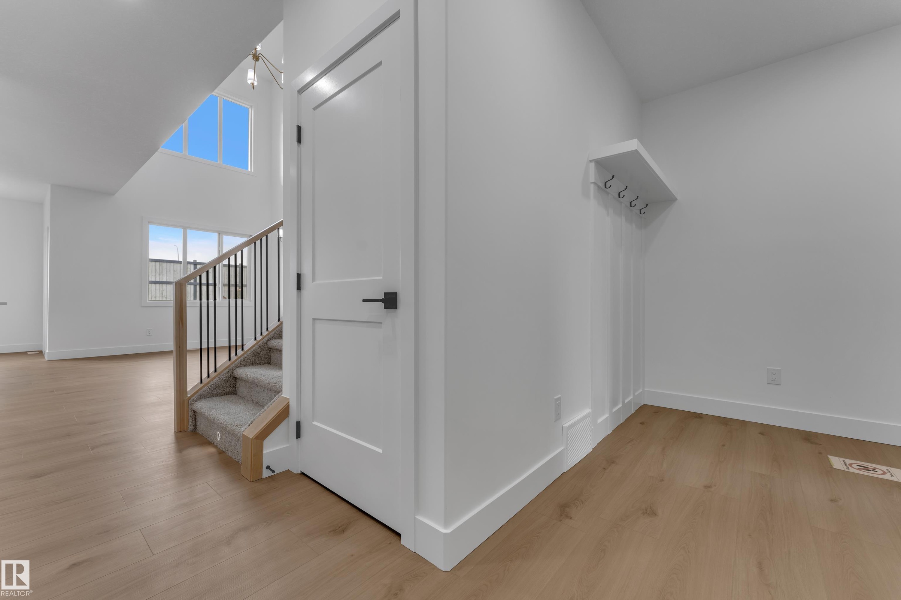 Wood-finish flooring throughout, white interior doors with contemporary black hardware, staircase with carpeted treads and metal spindles, built-in wall paneling detail, and high ceilings with upper-level windows - 178 Munn Way, Leduc, AB