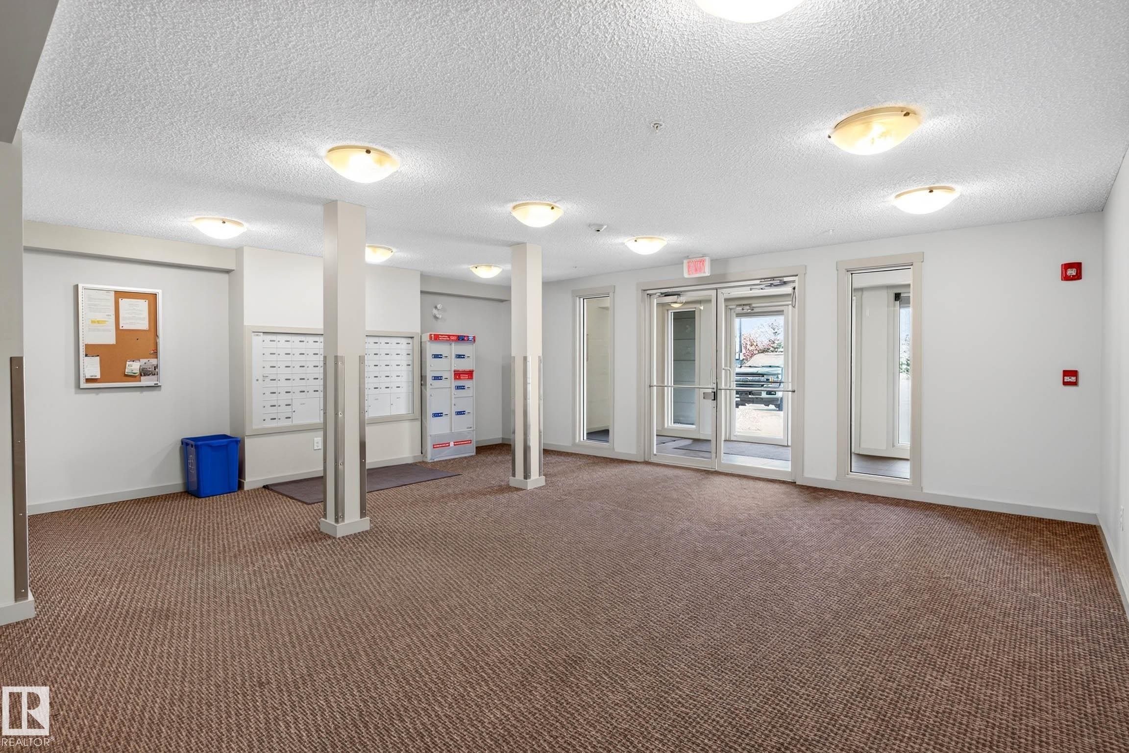 Spacious common area featuring textured carpet flooring, multiple ceiling-mounted light fixtures, and a glass-paneled entrance door - #408 25 Element Dr, St. Albert, AB