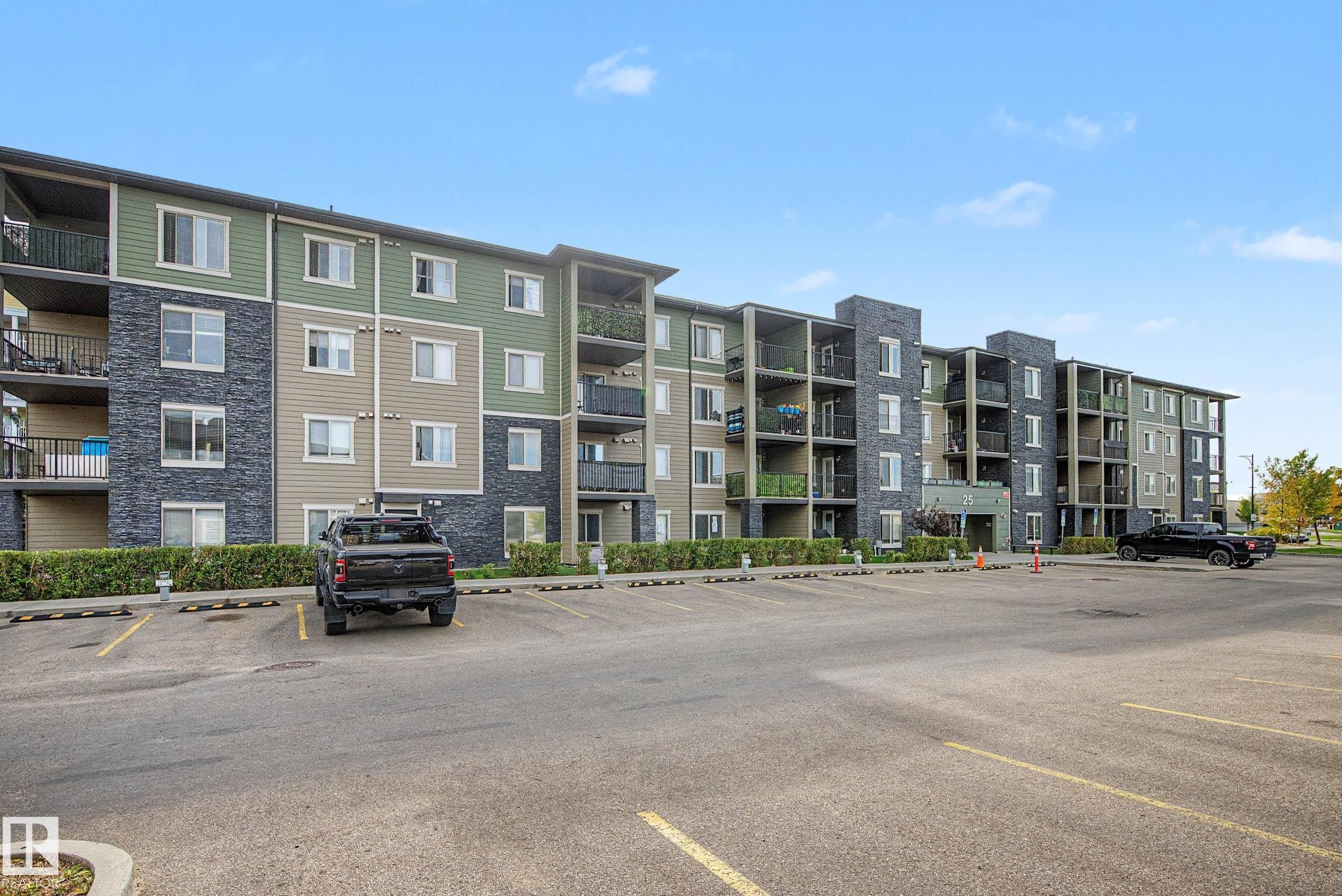 Multi-story building featuring a facade with mixed siding and stone accents, individual balconies, and an expansive paved parking lot - #408 25 Element Dr, St. Albert, AB
