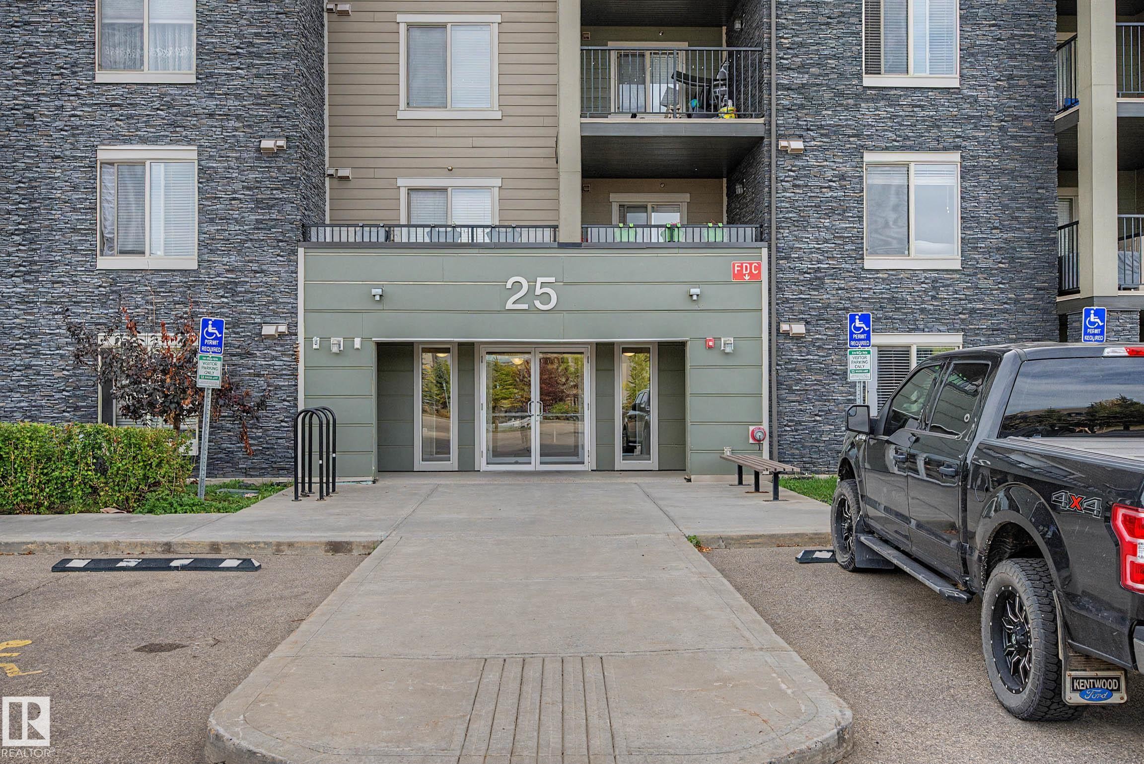 Building exterior with stone-look facade and multiple balconies, featuring a concrete walkway leading to a glass entrance - #408 25 Element Dr, St. Albert, AB
