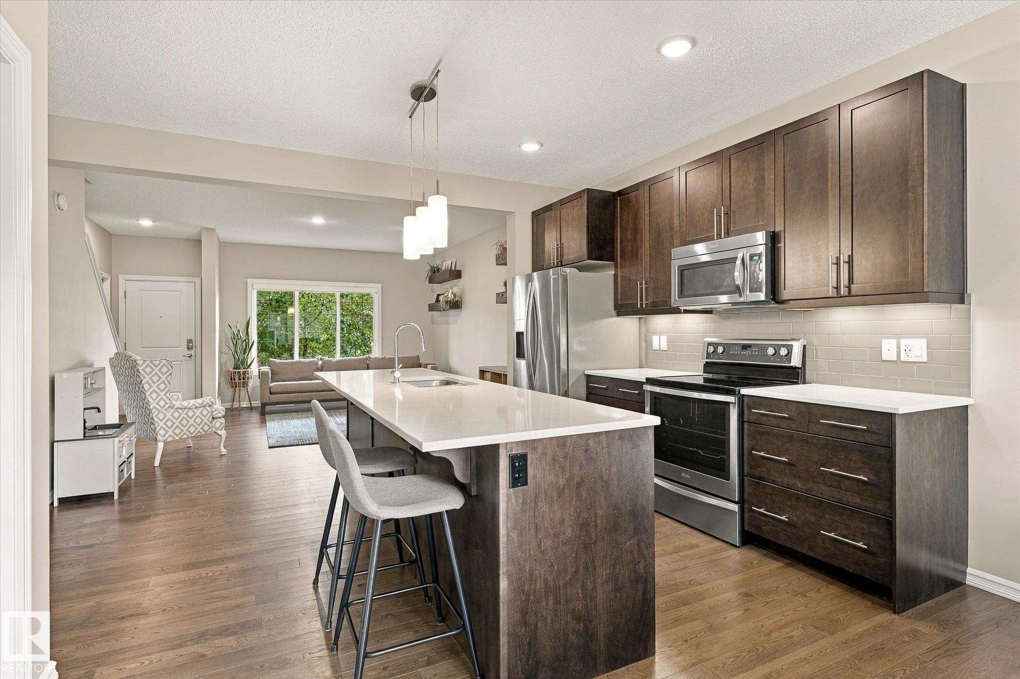 Open-concept kitchen featuring an eat-at island with a sink, wood-finish cabinetry, stainless steel appliances, and wood-finish flooring throughout - 21935 91 Avenue Nw, Edmonton, AB