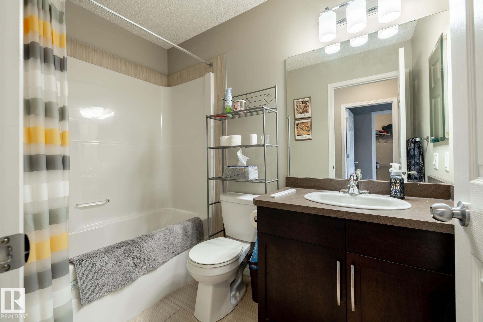 Bathroom featuring a dark-finish vanity with a stone-composite countertop, integrated oval sink, full-width vanity mirror, polished chrome lighting fixture, and a tub-shower combination with a surround - 2093 Chalmers Way Sw, Edmonton, AB