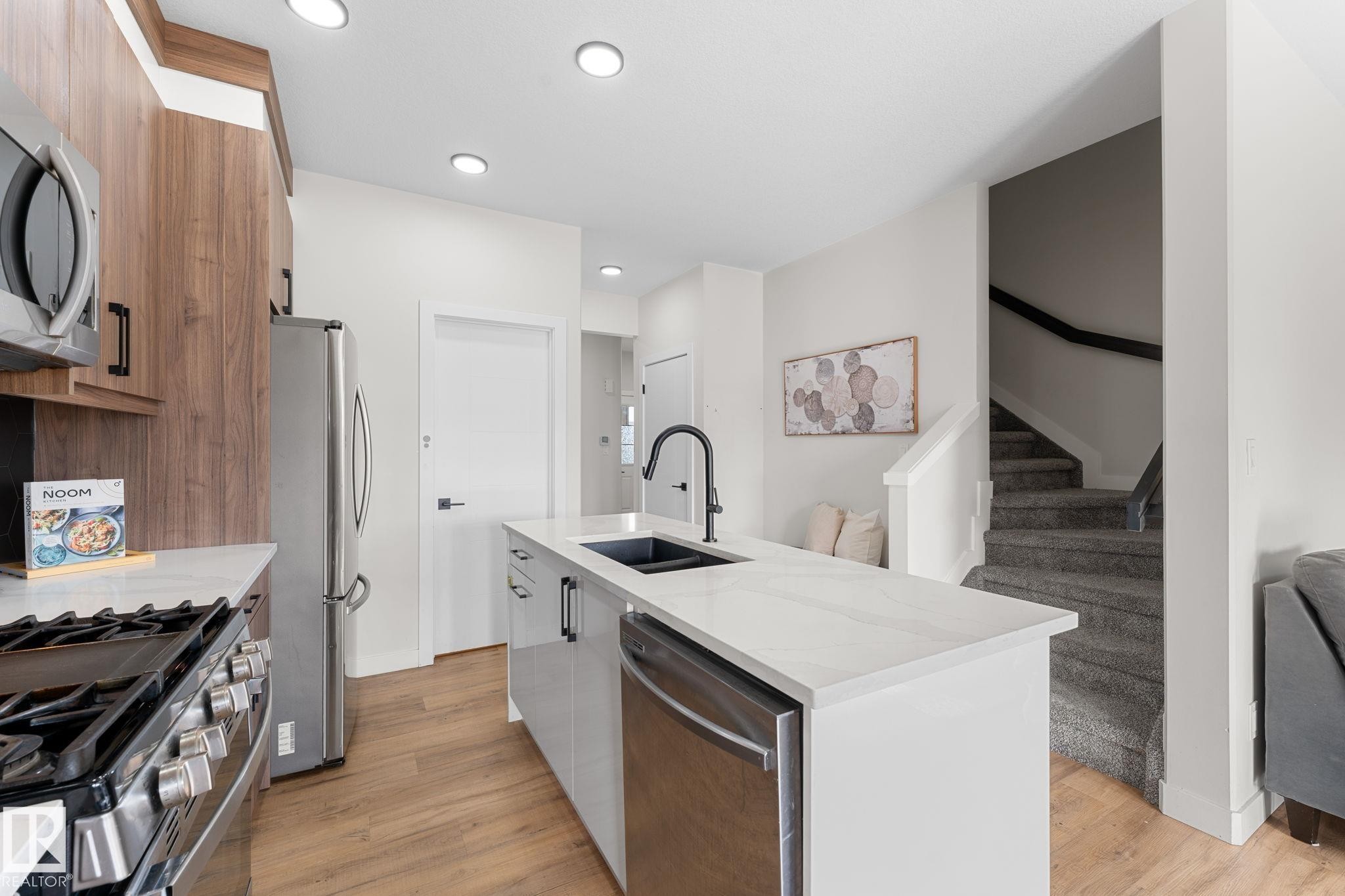 Modern kitchen featuring stainless steel appliances, a center island with a black sink and faucet, quartz countertops, and wood-finish flooring - 1142 Goldfinch Crescent Nw, Edmonton, AB