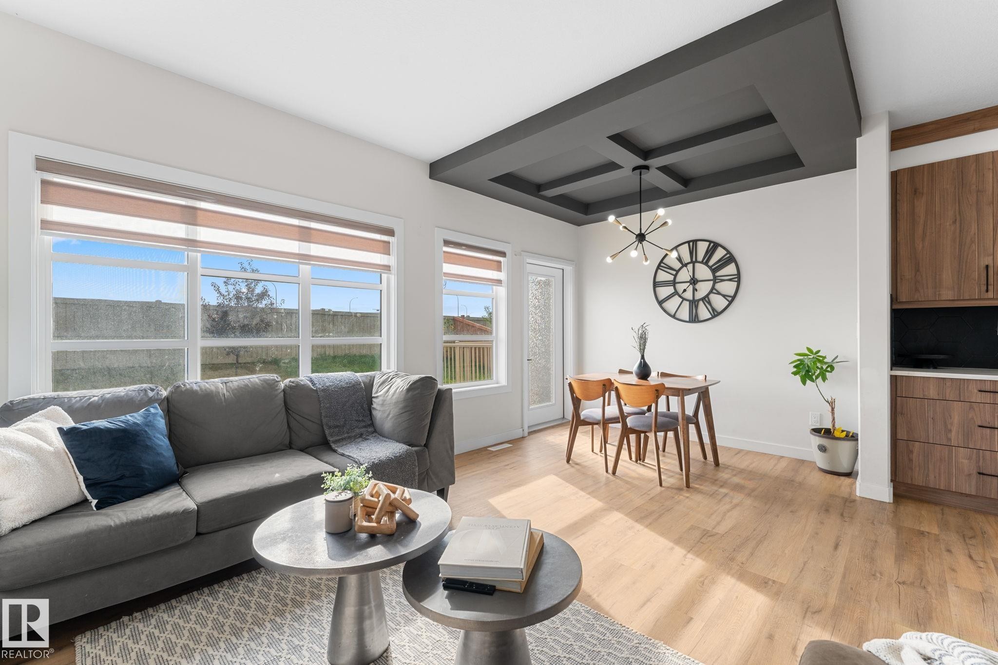 Wood-finish flooring throughout, coffered ceiling with a modern chandelier, large windows with roller shades, and built-in wood-finish cabinetry - 1142 Goldfinch Crescent Nw, Edmonton, AB