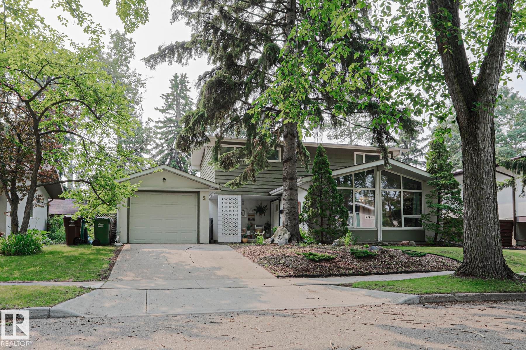 Two-story home featuring a single-car garage with an overhead door, a front-facing sunroom with expansive windows, and a landscaped front yard with mature trees - 5 Gilmore Crescent, St. Albert, AB