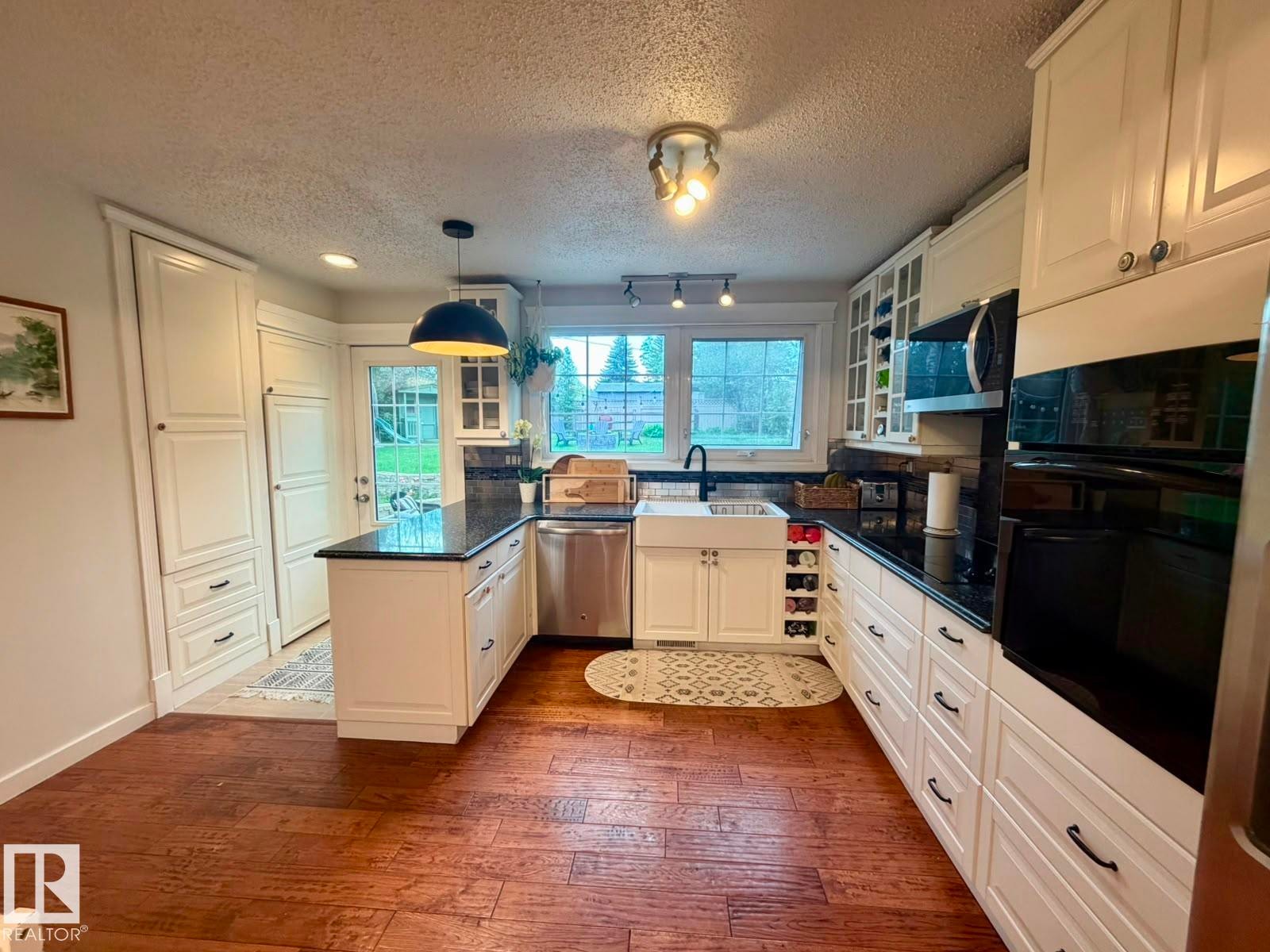Kitchen featuring white cabinetry, dark countertops, a farmhouse sink, stainless steel appliances, and wood-finish flooring - 5 Gilmore Crescent, St. Albert, AB