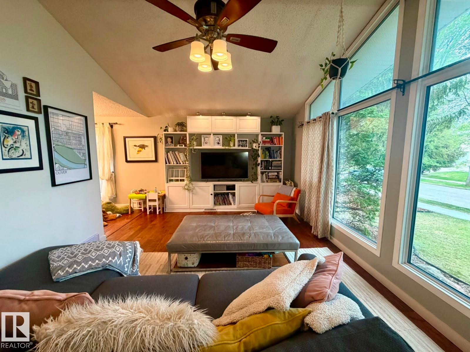 Vaulted ceiling, wood-finish flooring, white built-in media center with shelving, large picture windows, and a ceiling fan with integrated lighting - 5 Gilmore Crescent, St. Albert, AB