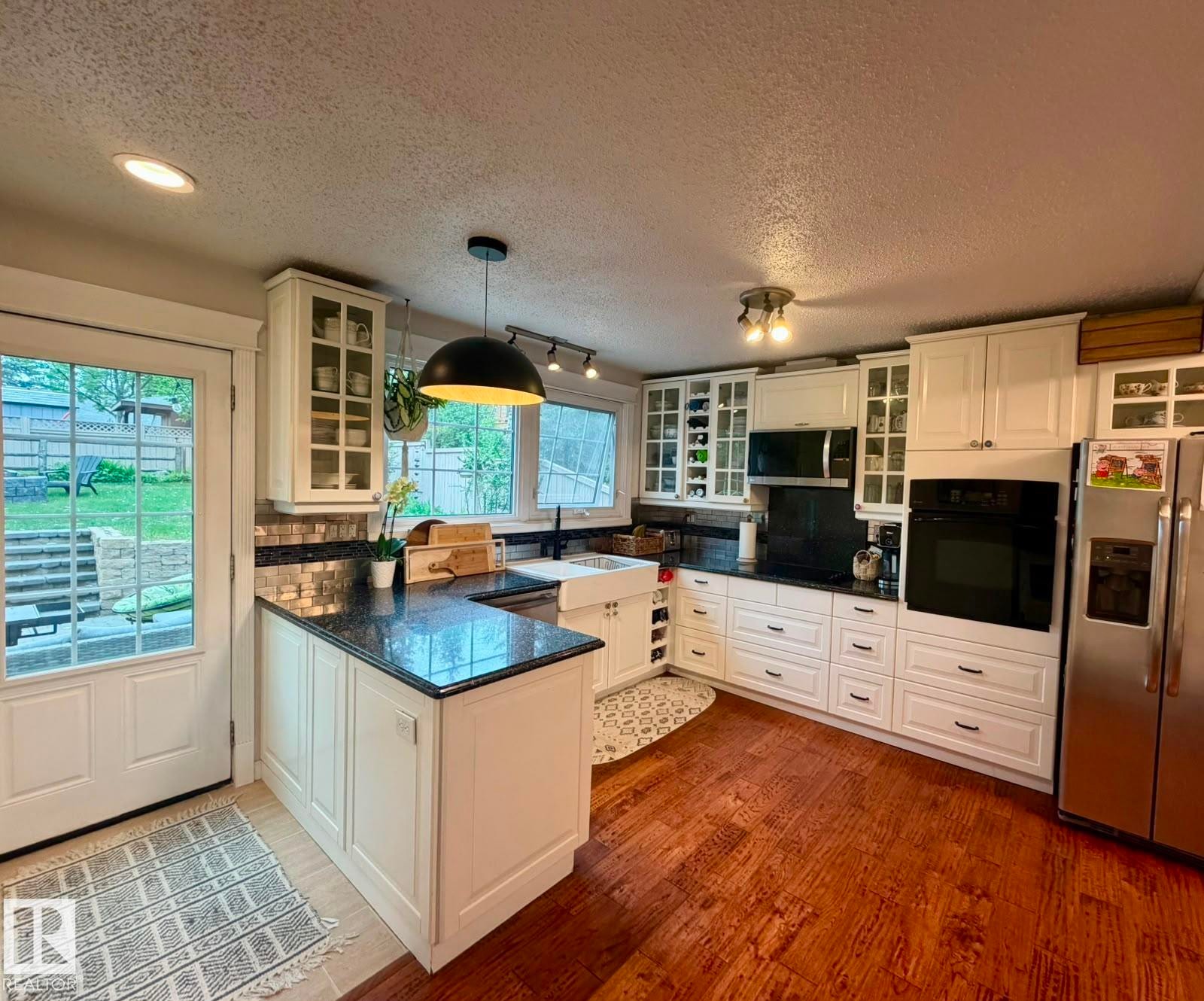 U-shaped kitchen with white cabinetry, dark stone countertops, wood-finish flooring, a white farmhouse sink, and stainless steel appliances - 5 Gilmore Crescent, St. Albert, AB