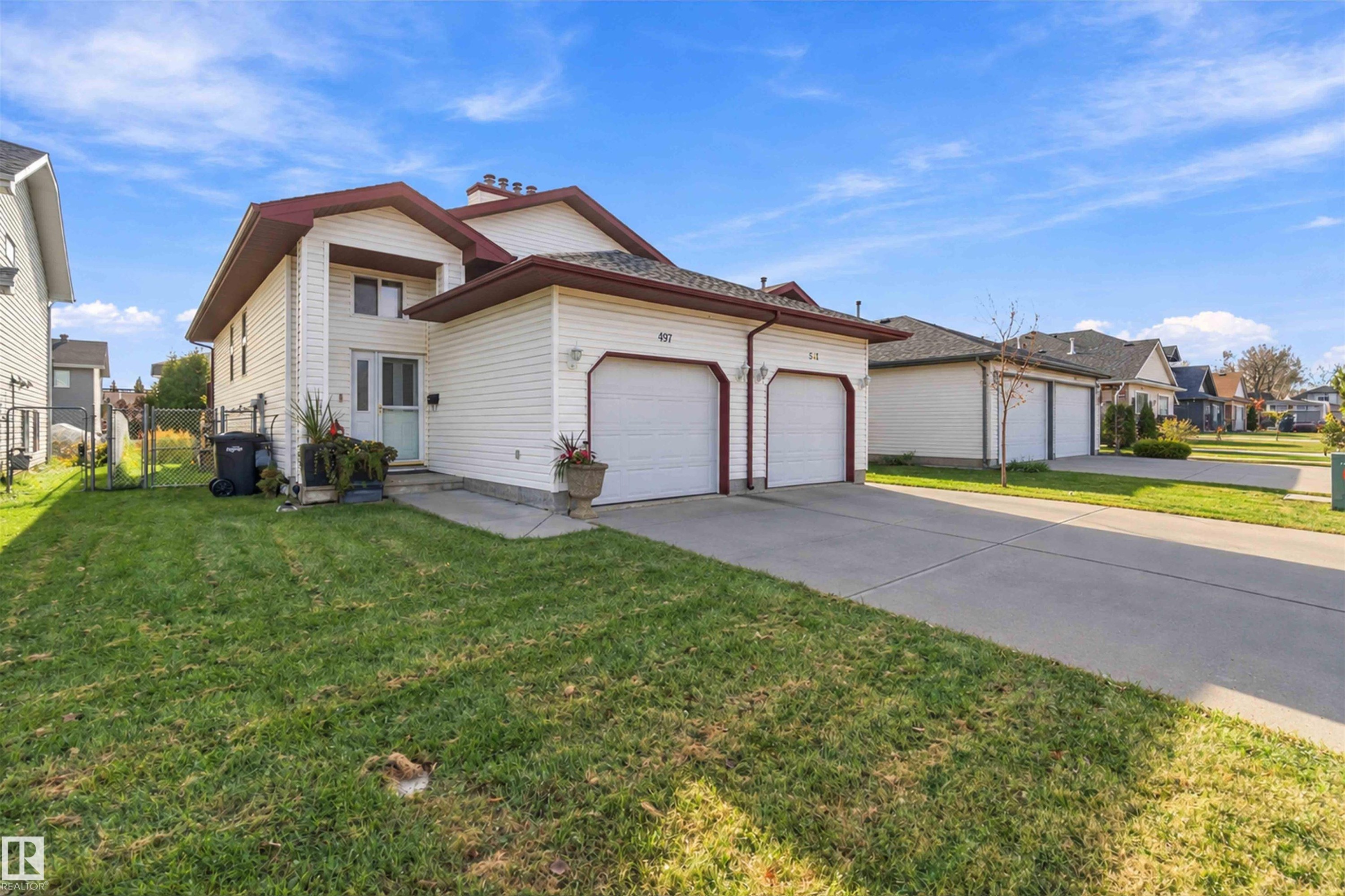 Two-story residence featuring light-toned vinyl siding, dual attached garages, a paved driveway, and a front-facing chimney - 497 Kananaskis Drive, Devon, AB