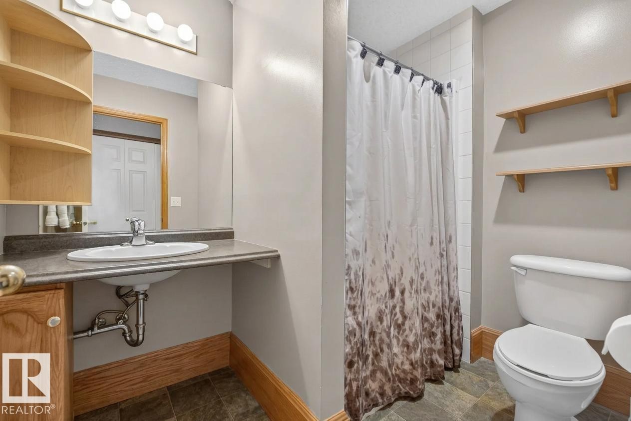 Bathroom featuring a vanity with an integrated basin, a large wall-mounted mirror, built-in corner shelving, stone-finish tile flooring, and wall-mounted wooden display shelves - 497 Kananaskis Drive, Devon, AB