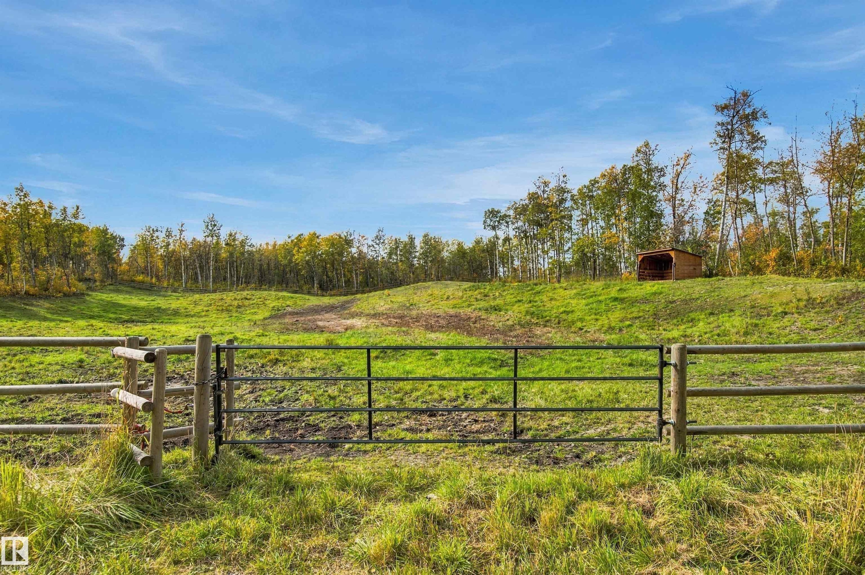 Expansive green pasture with a wood and metal gate, surrounded by mature trees, and featuring a small shelter on the hillside - 51064 Rge Rd 222, Rural Strathcona County, AB