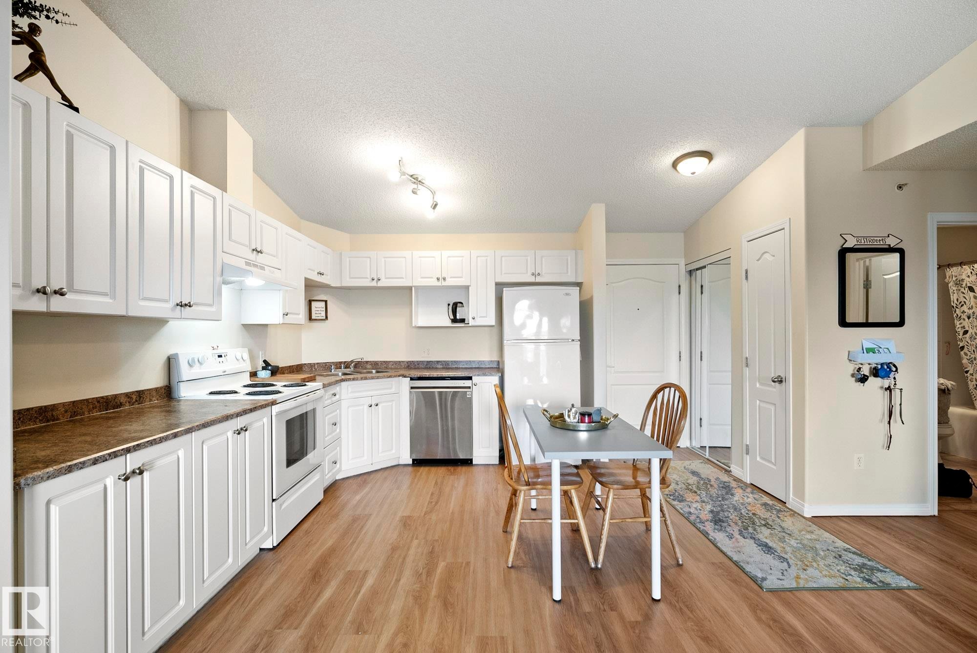 Kitchen featuring white cabinetry, wood-finish flooring, a stainless steel dishwasher, and built-in overhead lighting - #416 111 Edwards Dr Sw, Edmonton, AB