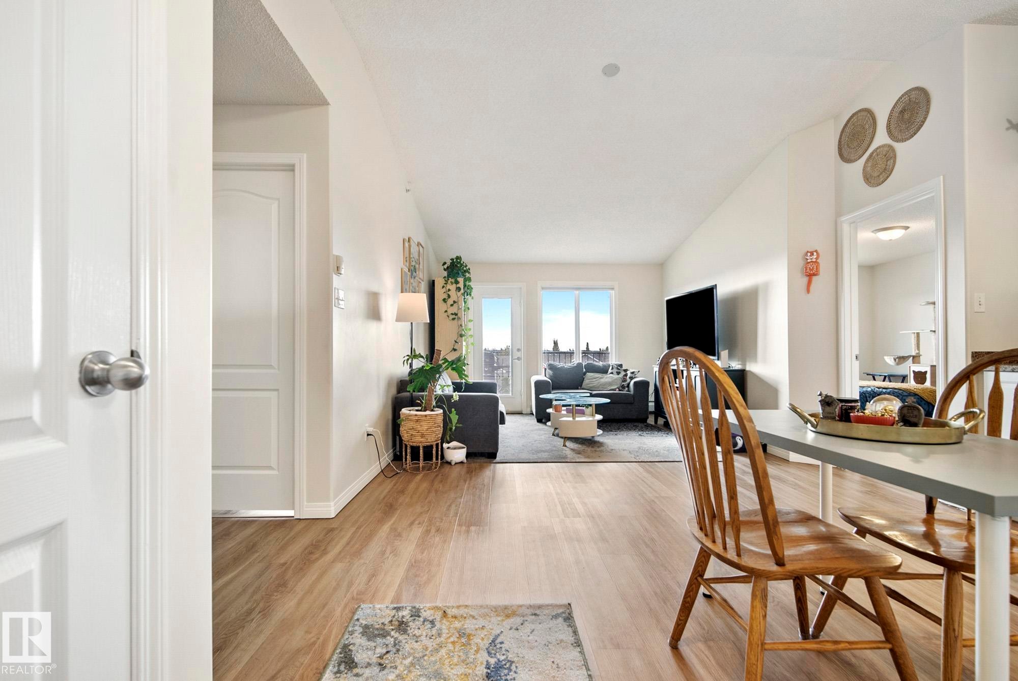 Open-concept living area with wood-finish flooring, vaulted ceilings, and a large glass door leading to an exterior balcony - #416 111 Edwards Dr Sw, Edmonton, AB