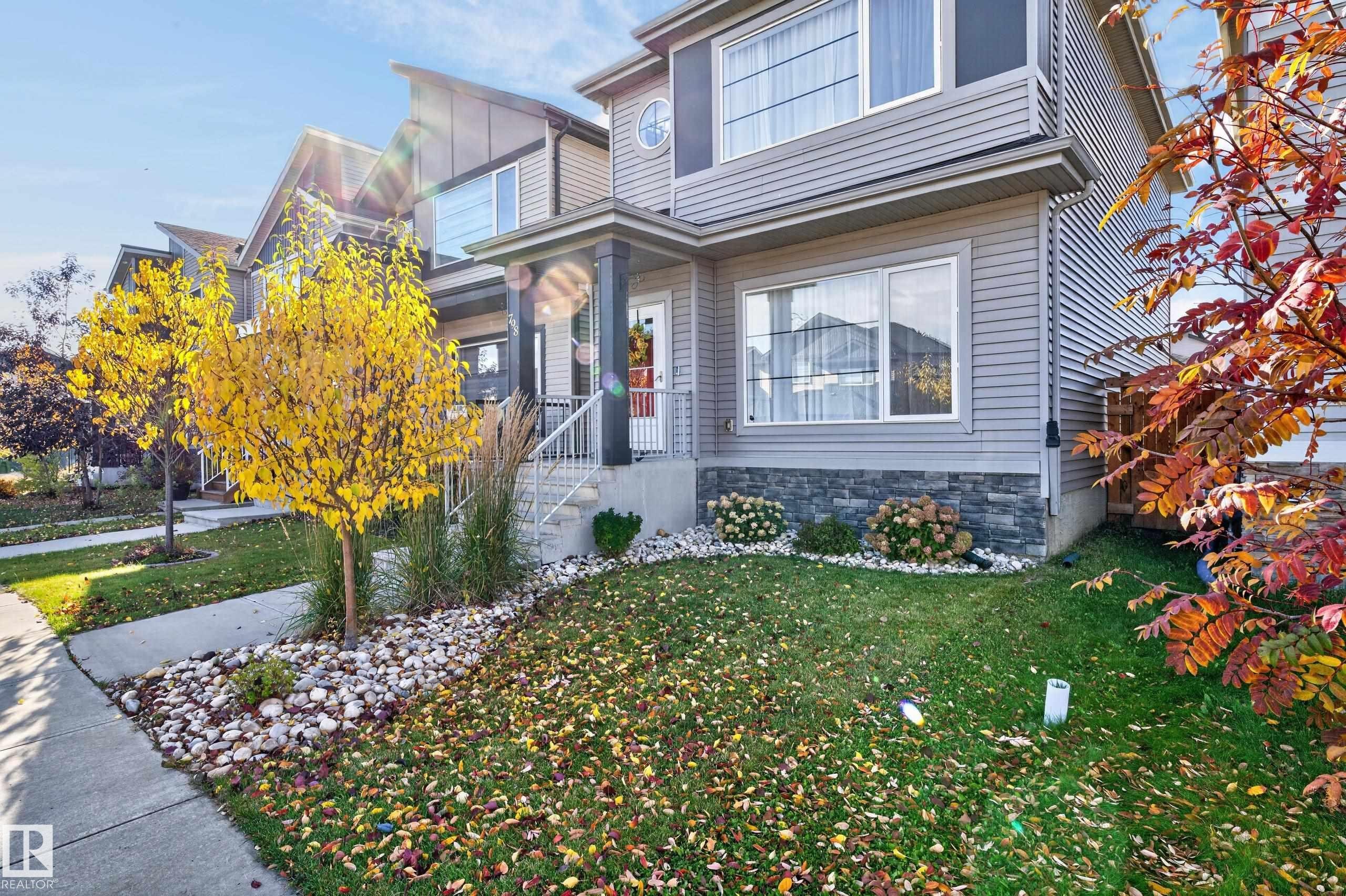 Two-story exterior featuring light gray horizontal siding, stone veneer masonry, a covered porch with metal railing, and an architectural window with a circular accent - 798 Berg Loop, Leduc, AB