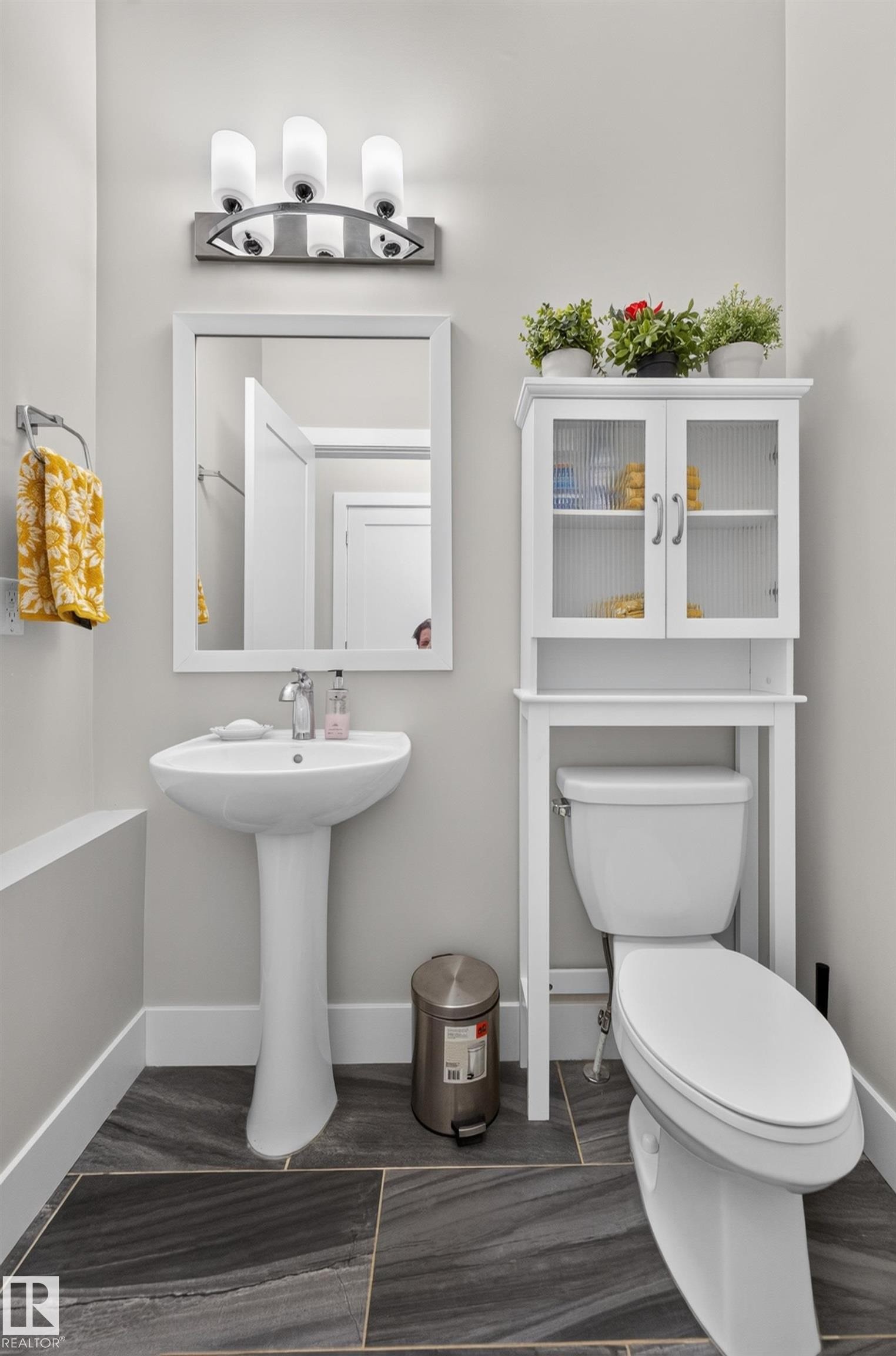 Pedestal sink with polished chrome faucet, three-light vanity fixture, rectangular framed mirror, and large-format tile flooring - 798 Berg Loop, Leduc, AB