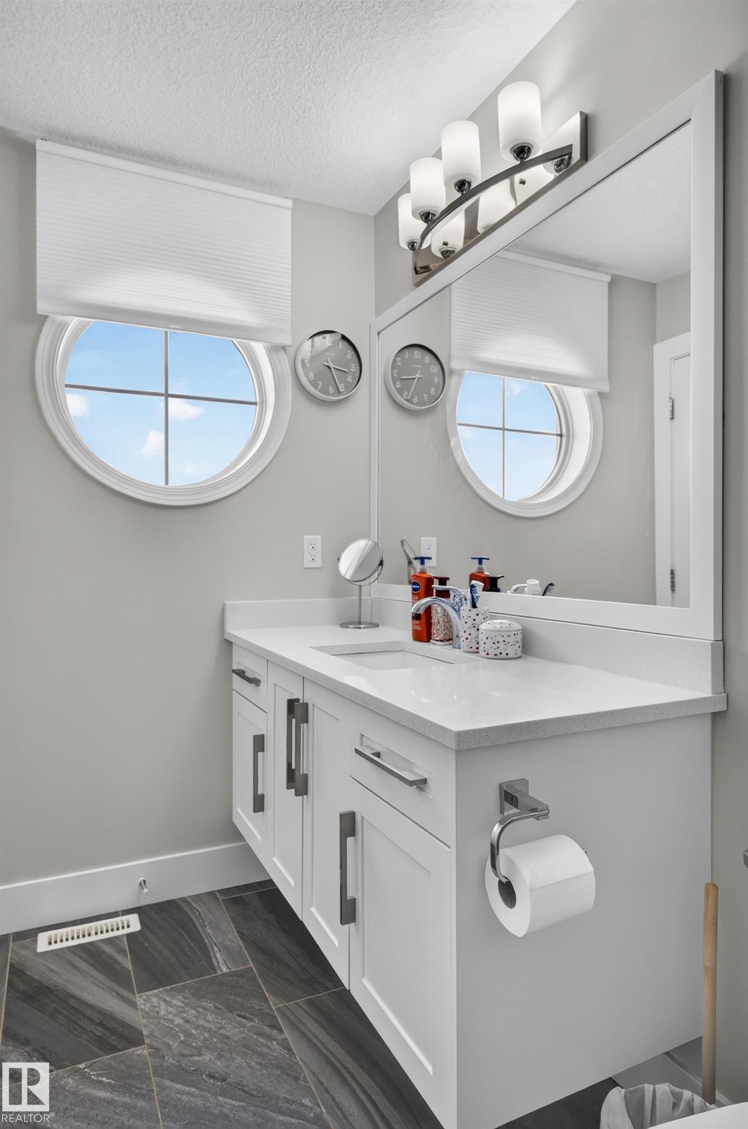 White vanity with a solid-surface countertop and integrated sink, framed rectangular wall mirror, circular window, and charcoal-toned tile flooring - 798 Berg Loop, Leduc, AB