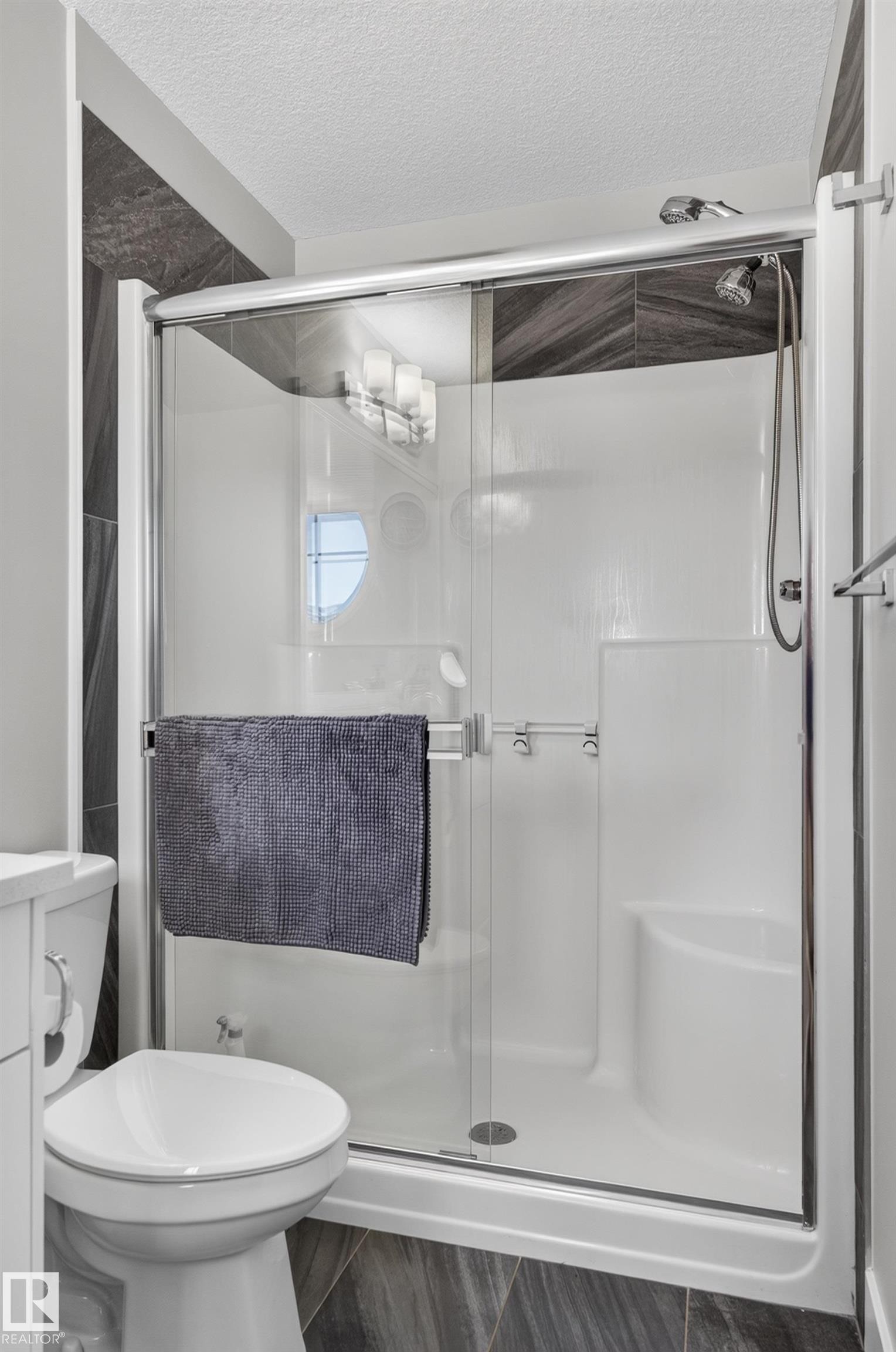 Shower enclosure featuring sliding glass doors, a built-in corner seat, and a handheld showerhead, accented by dark, chevron-patterned tile surrounds and wood-finish floor tiling - 798 Berg Loop, Leduc, AB