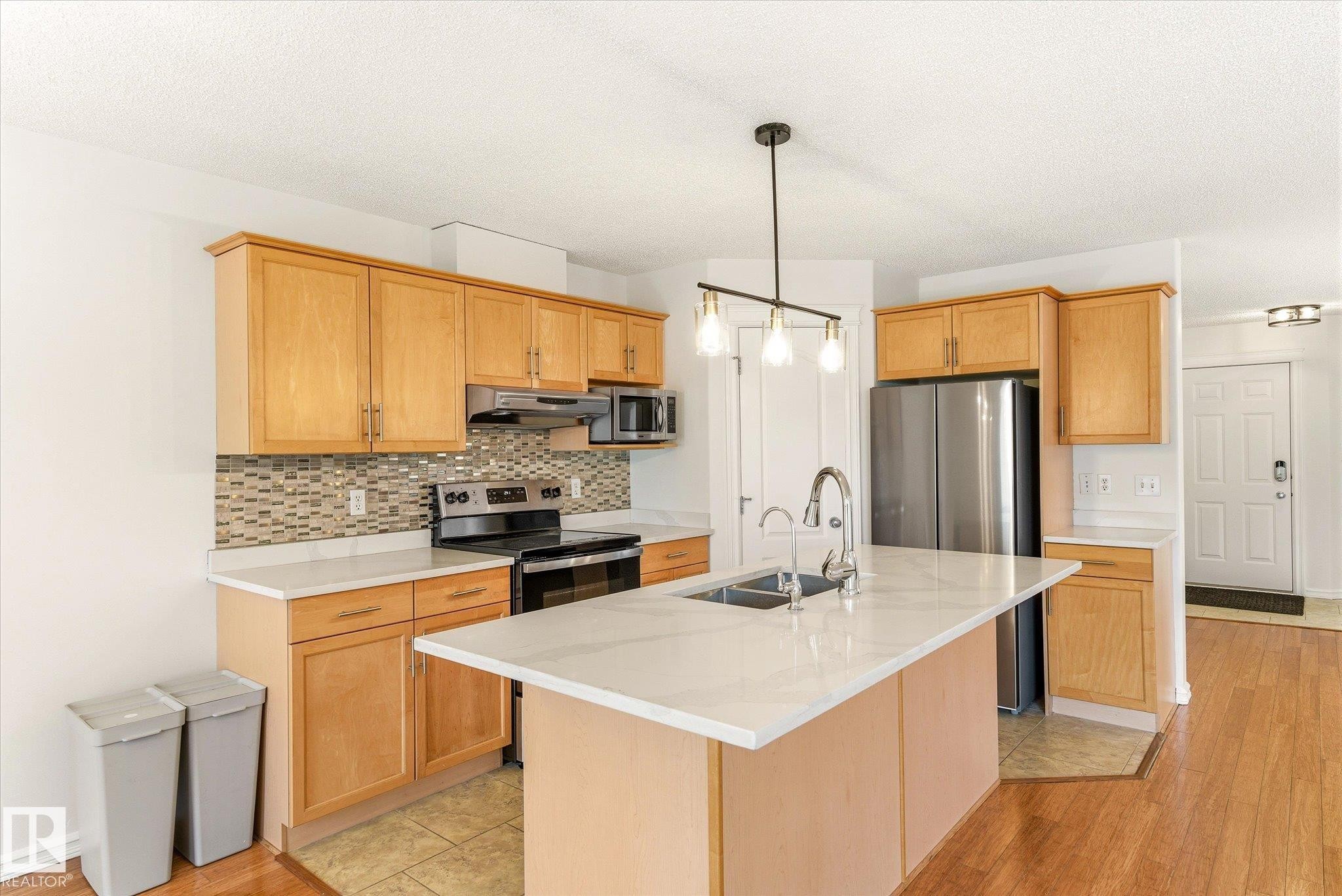 Kitchen featuring light wood-finish cabinetry, stainless steel appliances, a mosaic tile backsplash, and an island with an integrated sink and pendant lighting - 2550 Rabbit Hill Road Nw, Edmonton, AB