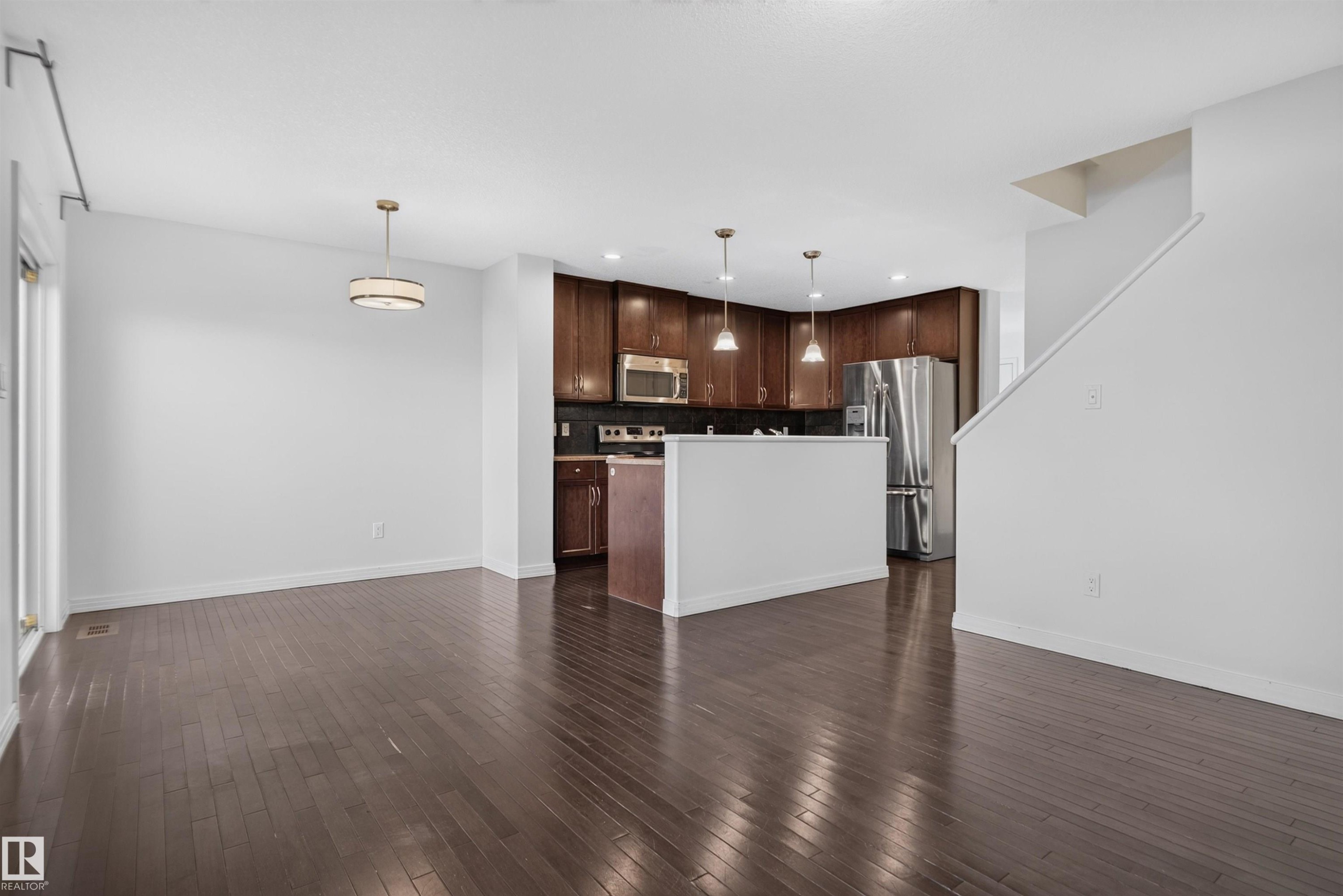 Expansive wood-finish flooring throughout, featuring a kitchen with dark wood cabinetry, stainless steel appliances, and pendant lighting - 2145 28 Street Nw, Edmonton, AB
