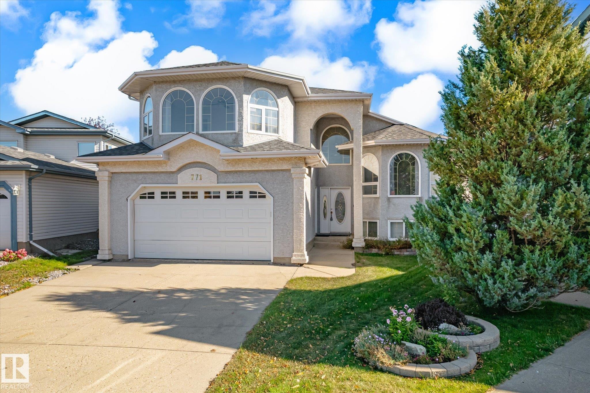 Two-story stucco exterior with decorative arched windows, an attached two-car garage, concrete driveway, and mature landscaping - 771 Johns Road Nw, Edmonton, AB