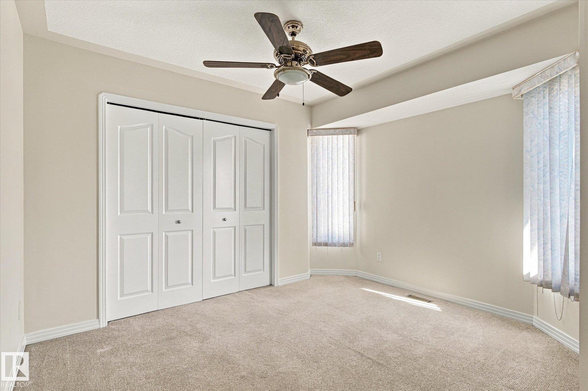 Carpeted room featuring a ceiling fan, dual bi-fold closet doors, and bay windows with vertical blinds - 771 Johns Road Nw, Edmonton, AB