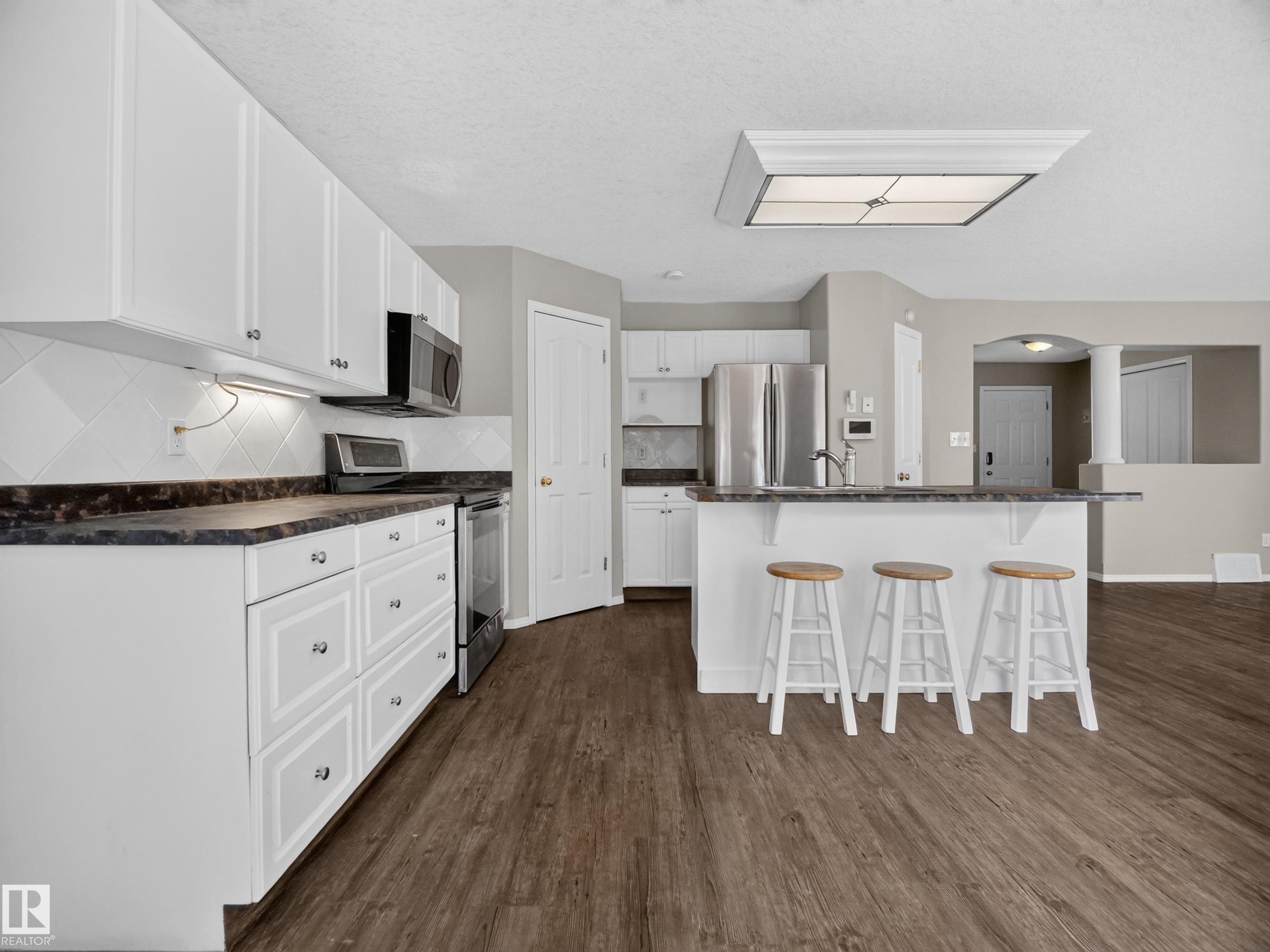 Kitchen featuring white cabinetry, dark countertops, a breakfast bar, stainless steel appliances, and wood-finish flooring - 14 Wells Court, Leduc, AB