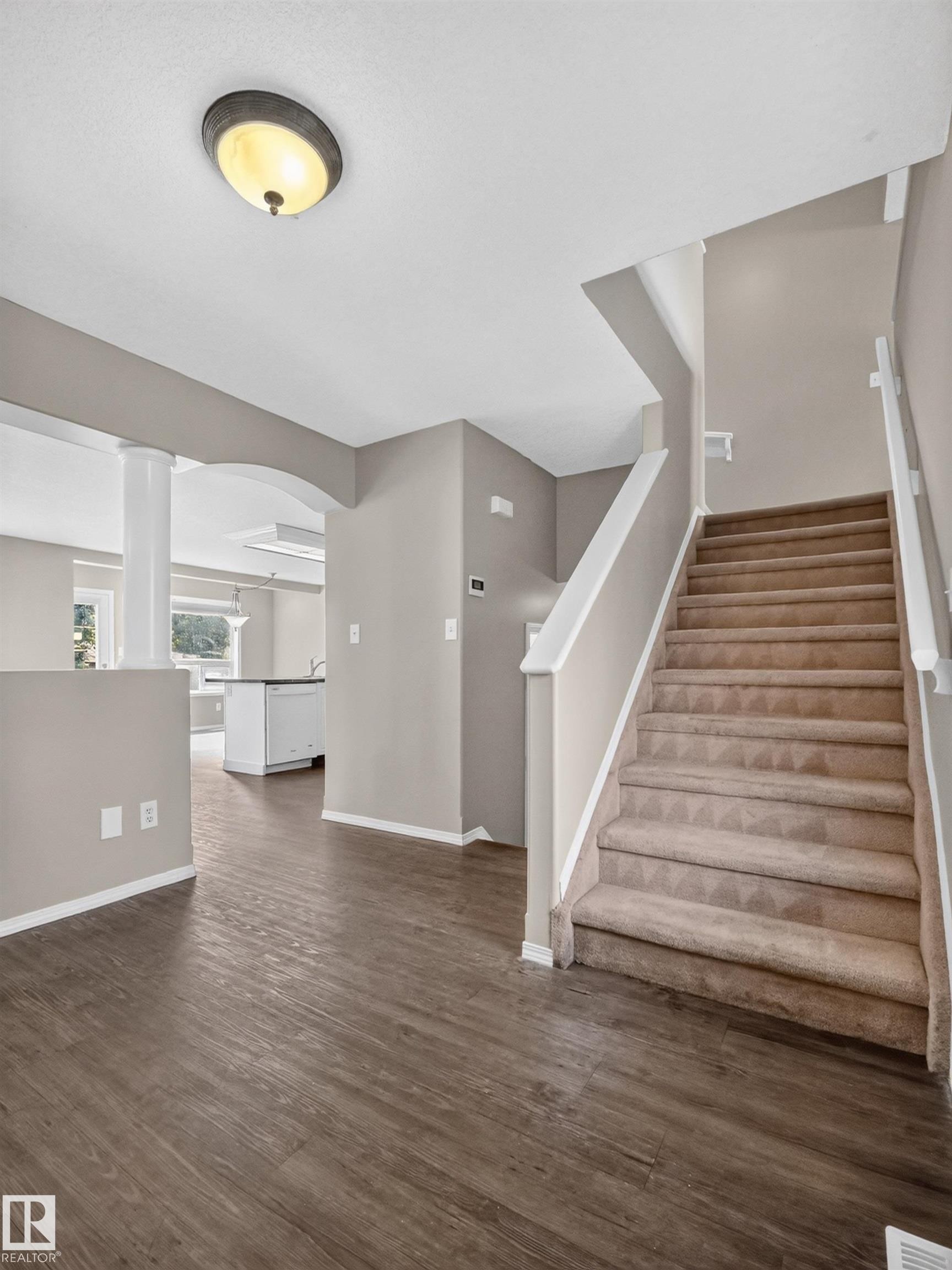 Wood-finish flooring, staircase with carpeting, recessed ceiling light, white trim, and a partial view of a kitchen with white cabinetry - 14 Wells Court, Leduc, AB