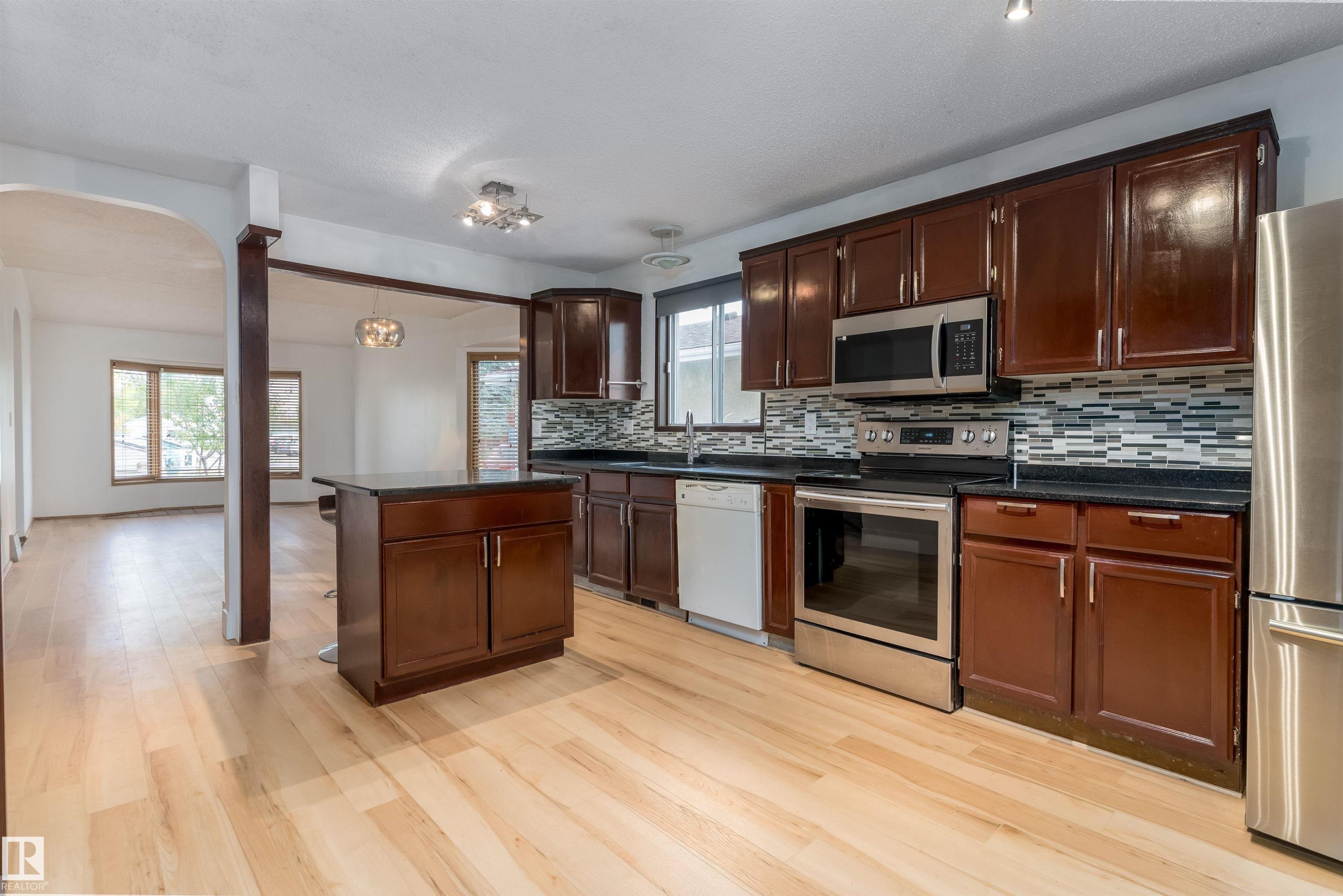 Kitchen featuring wood-finish flooring, dark wood cabinetry, a central island, stainless steel appliances, and a mosaic tile backsplash - 18739 72 Avenue Nw, Edmonton, AB