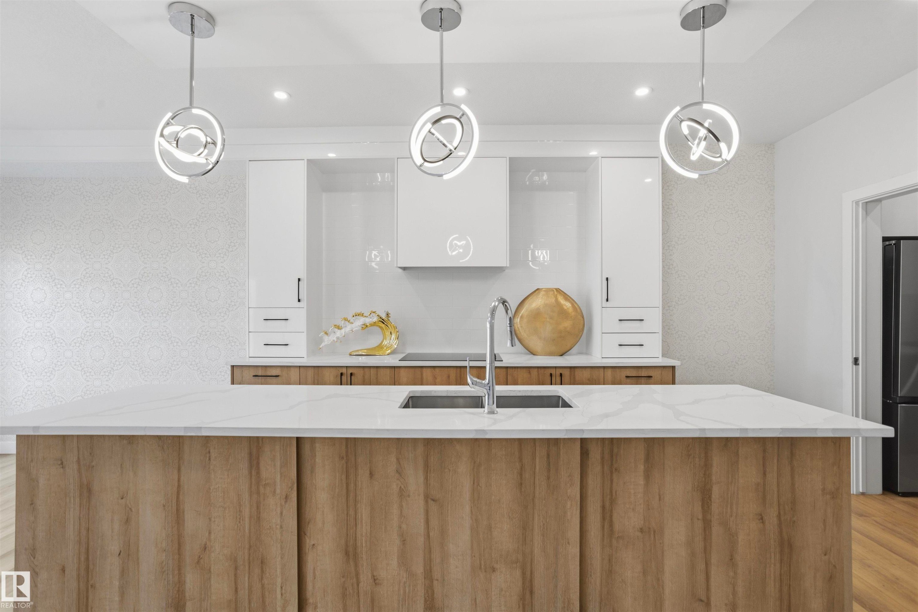 Kitchen featuring a large central island with light-toned wood-finish cabinetry and a quartz countertop, complemented by white cabinetry, a built-in range hood, and three modern spherical pendant lights - 2045 209A St Nw, Edmonton, AB