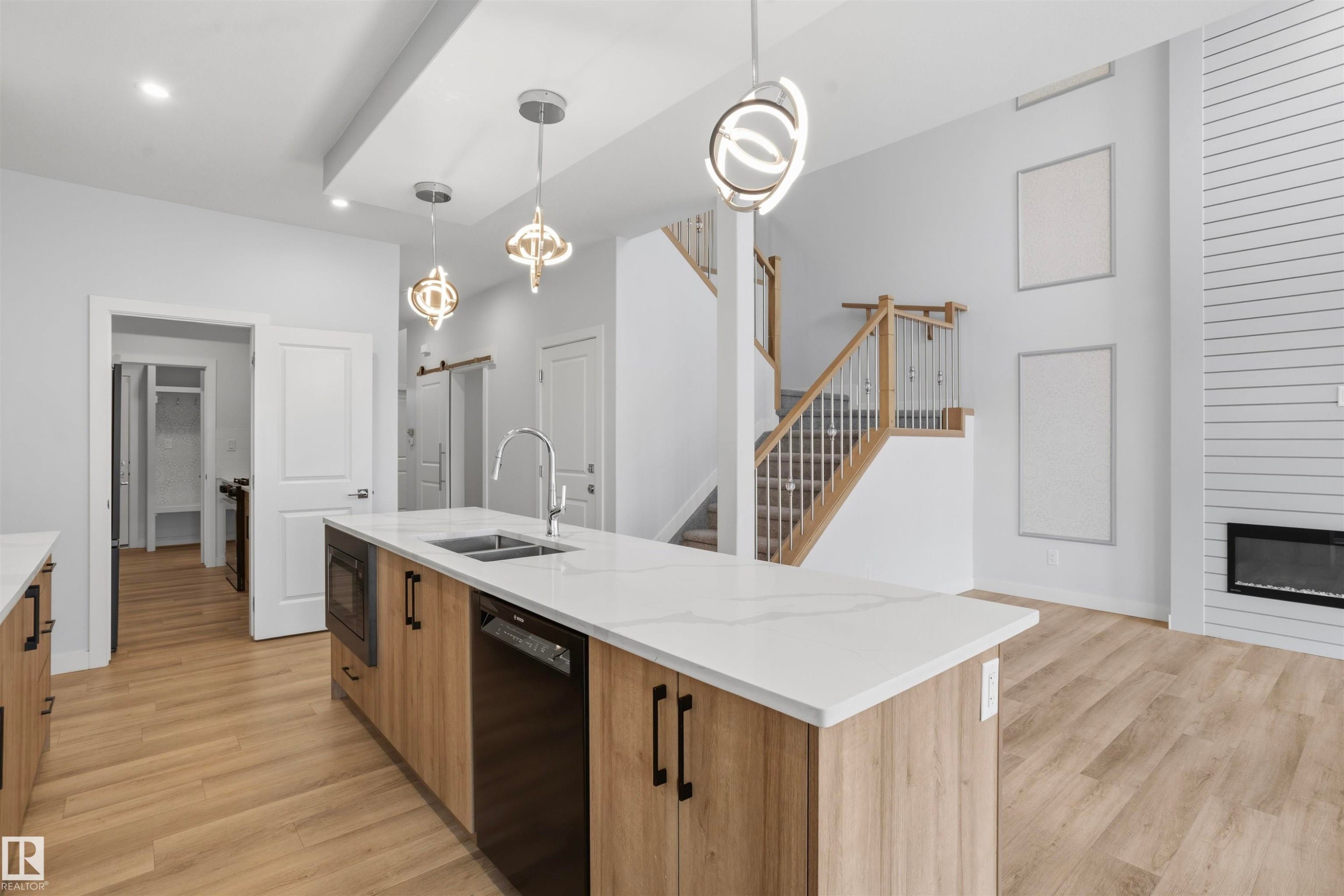 Kitchen island with a light-toned wood finish, quartz countertop, and undermount dual-basin sink, complemented by three spherical pendant lights, wood-finish flooring, and a fireplace with shiplap surround - 2045 209A St Nw, Edmonton, AB