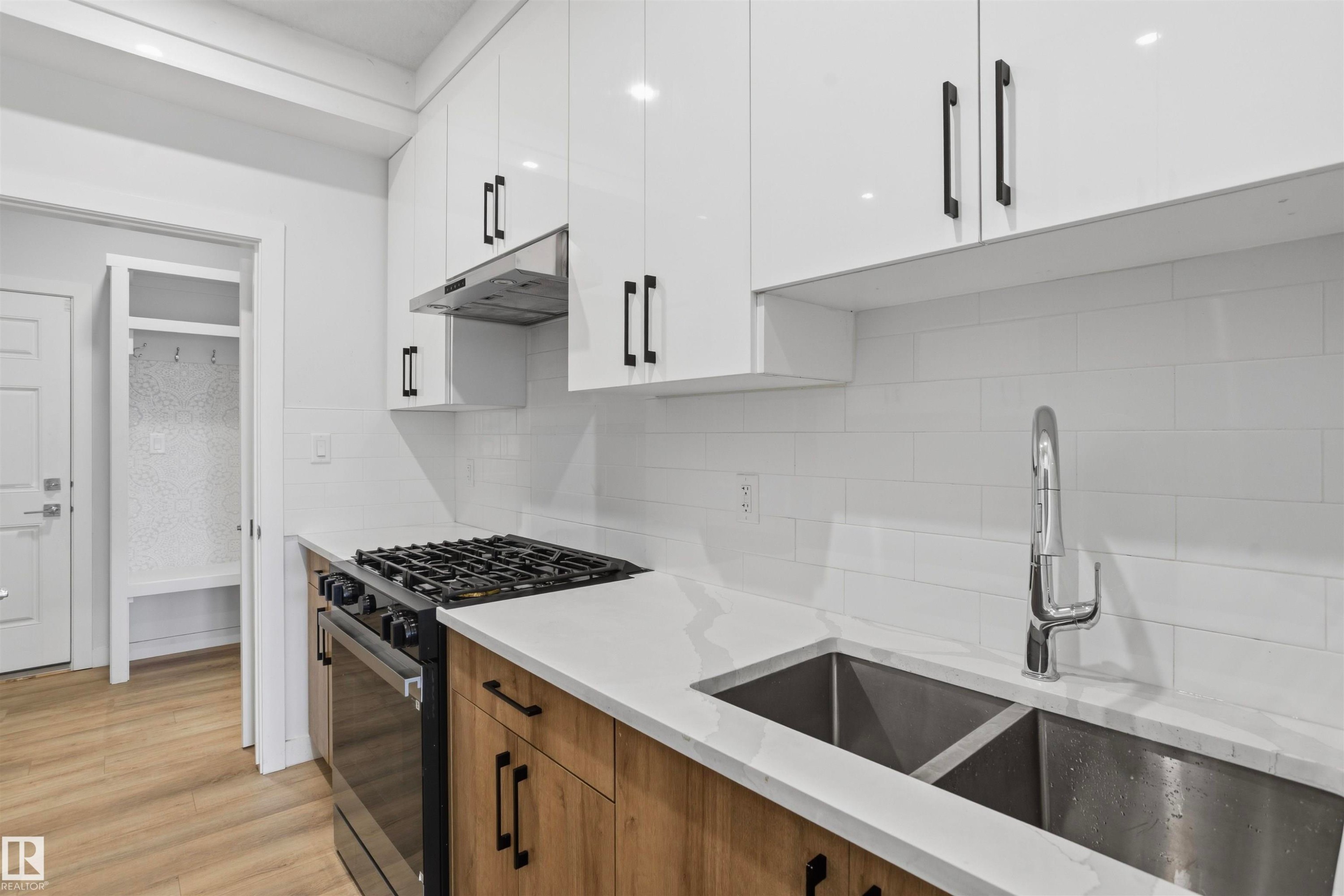 Modern kitchen featuring high-gloss white upper cabinetry, wood-finish lower cabinetry, stone-style countertops, white subway tile backsplash, and stainless steel integrated double-basin sink with pull-down faucet - 2045 209A St Nw, Edmonton, AB