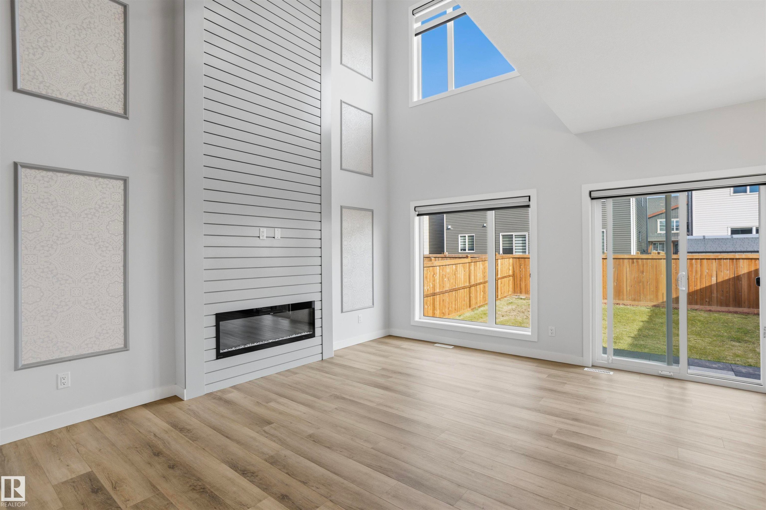 Double-height ceiling, horizontal slat-accented fireplace wall, floor-to-ceiling windows, wood-finish flooring, and glass sliding door - 2045 209A St Nw, Edmonton, AB